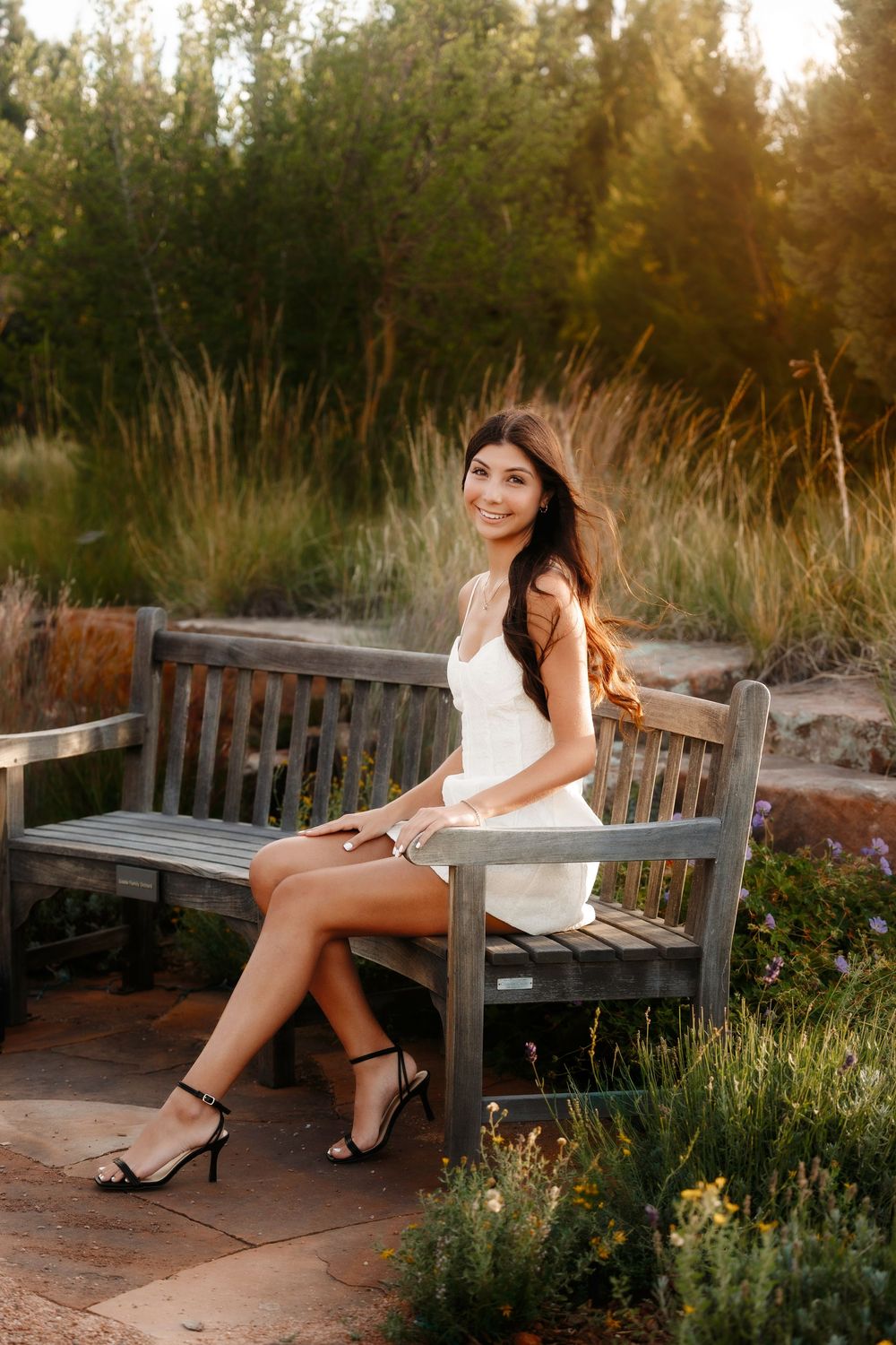 Elegant young woman in white dress sitting on wooden bench surrounded by lush greenery at sunset.