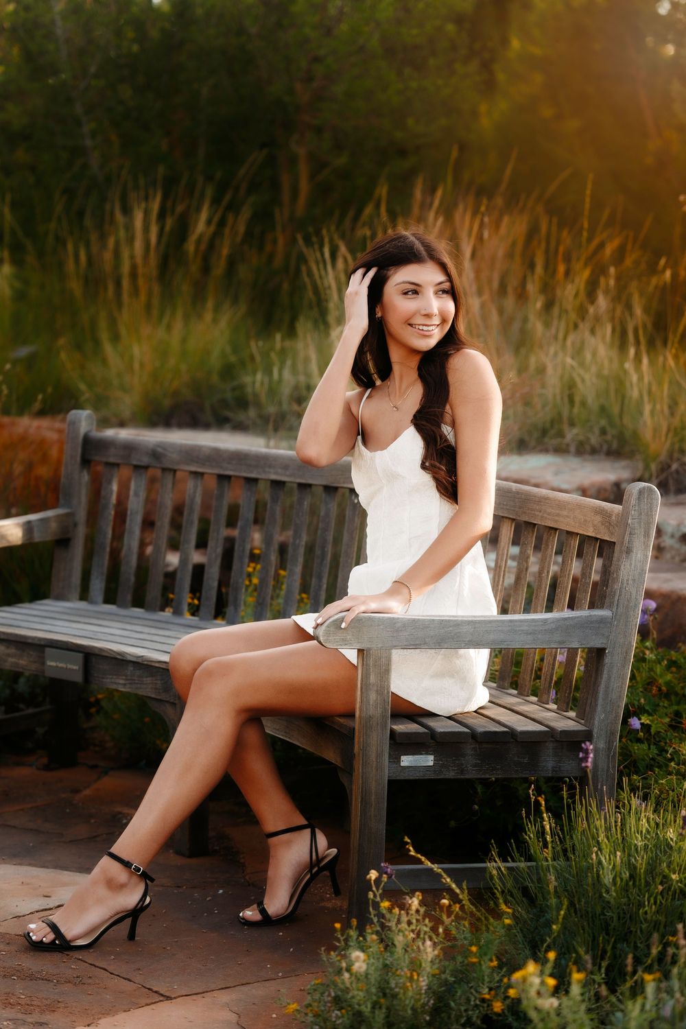 Young woman in white dress sitting on a wooden bench in a sunlit garden setting.