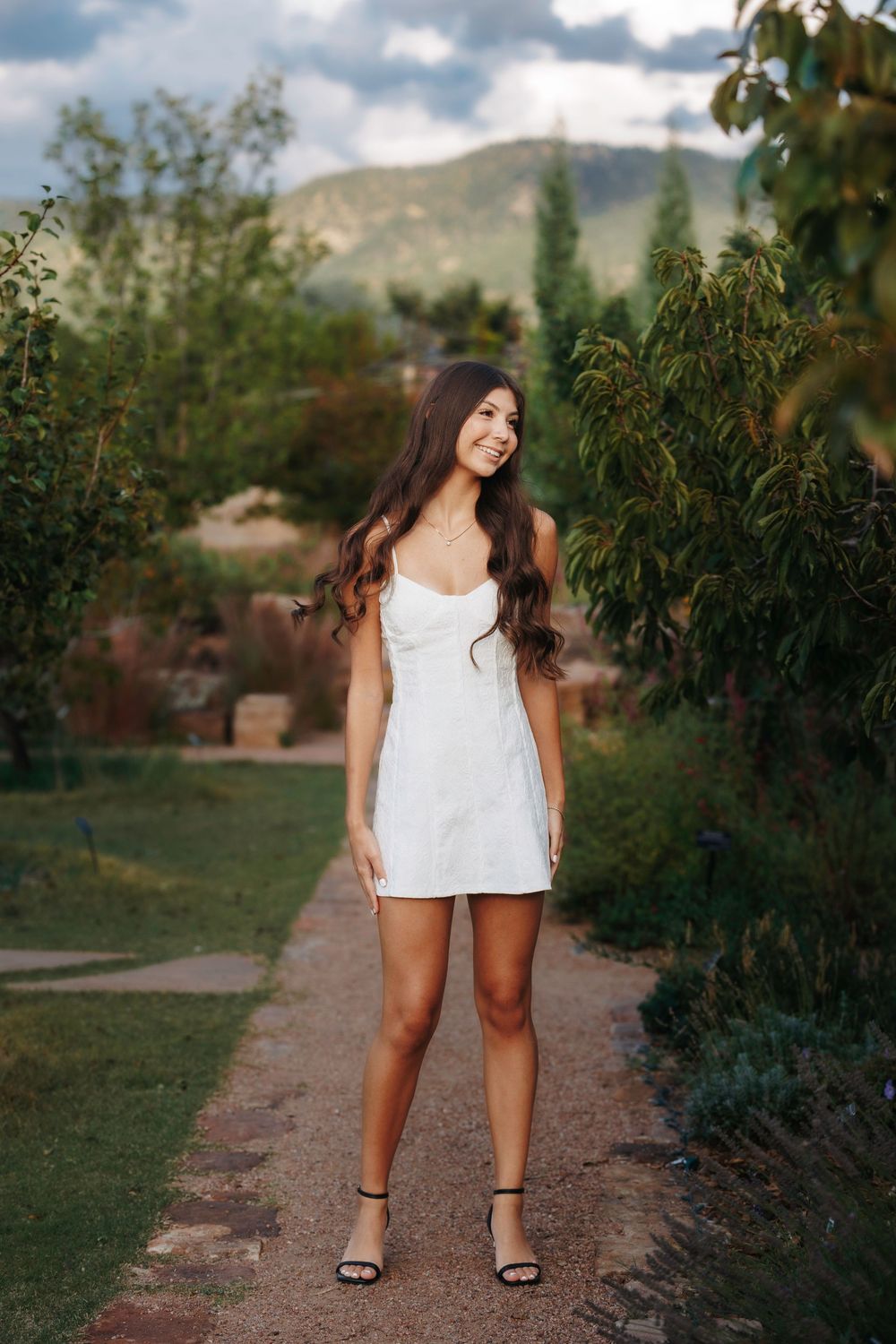 Smiling brunette woman in white mini dress standing on garden path with mountains and cypress trees behind her.