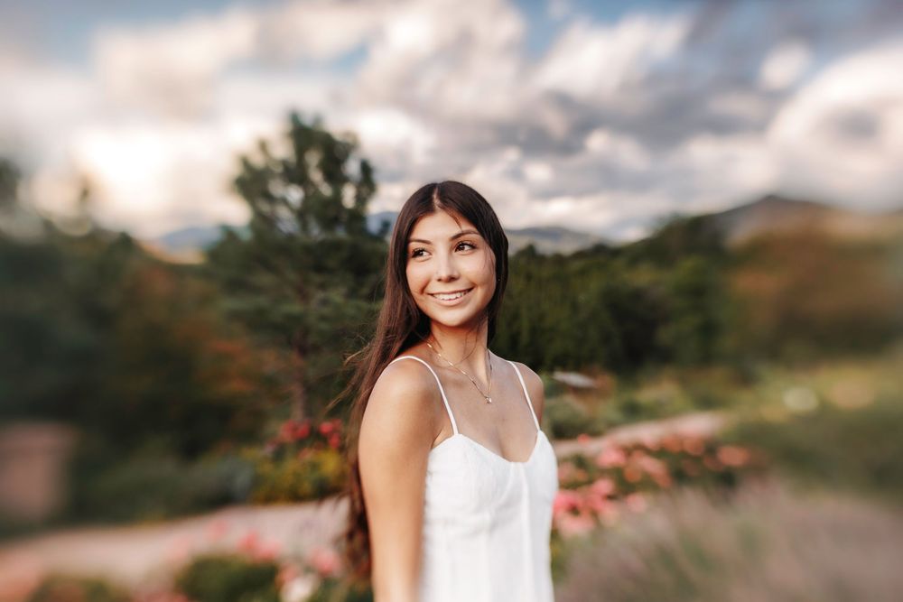 Smiling young woman in white sundress with soft bokeh garden and mountain background on a cloudy day.
