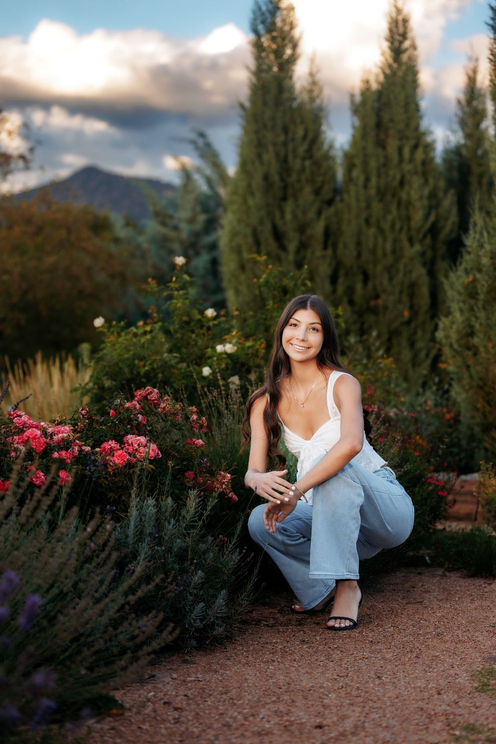 Santa Fe senior in white top and jeans crouching beside blooming roses and lavender with cypress trees behind her.