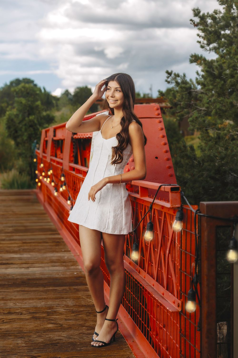 Young woman in white dress leaning on a red bridge railing with bokeh string lights.