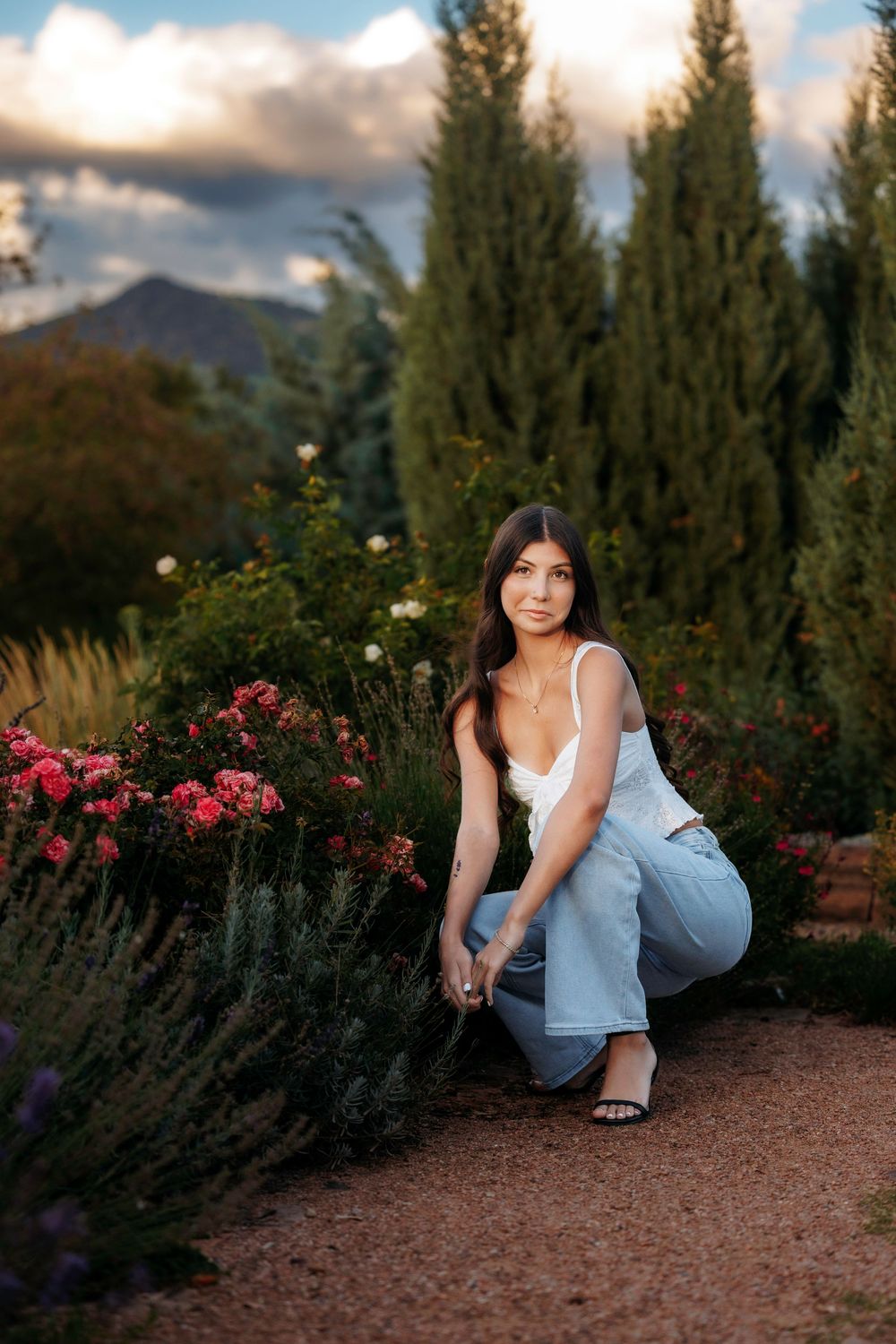 Santa Fe senior in white tank and jeans crouching on garden path surrounded by roses and tall cypress trees.