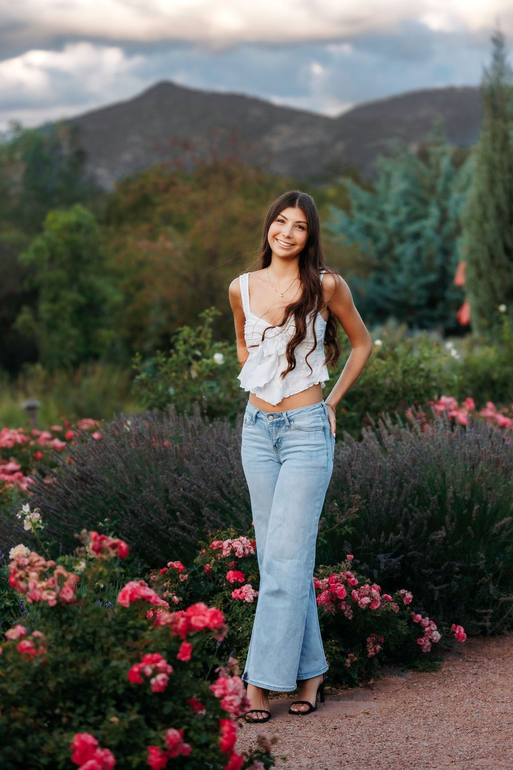 Santa Fe senior in white ruffled crop top and flare jeans posing among pink roses with mountain backdrop.