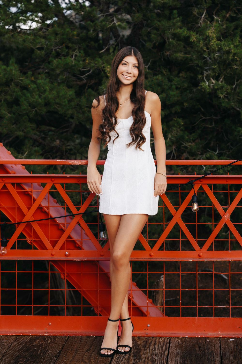 Smiling teenage girl in white mini dress posing on a red bridge with string lights.