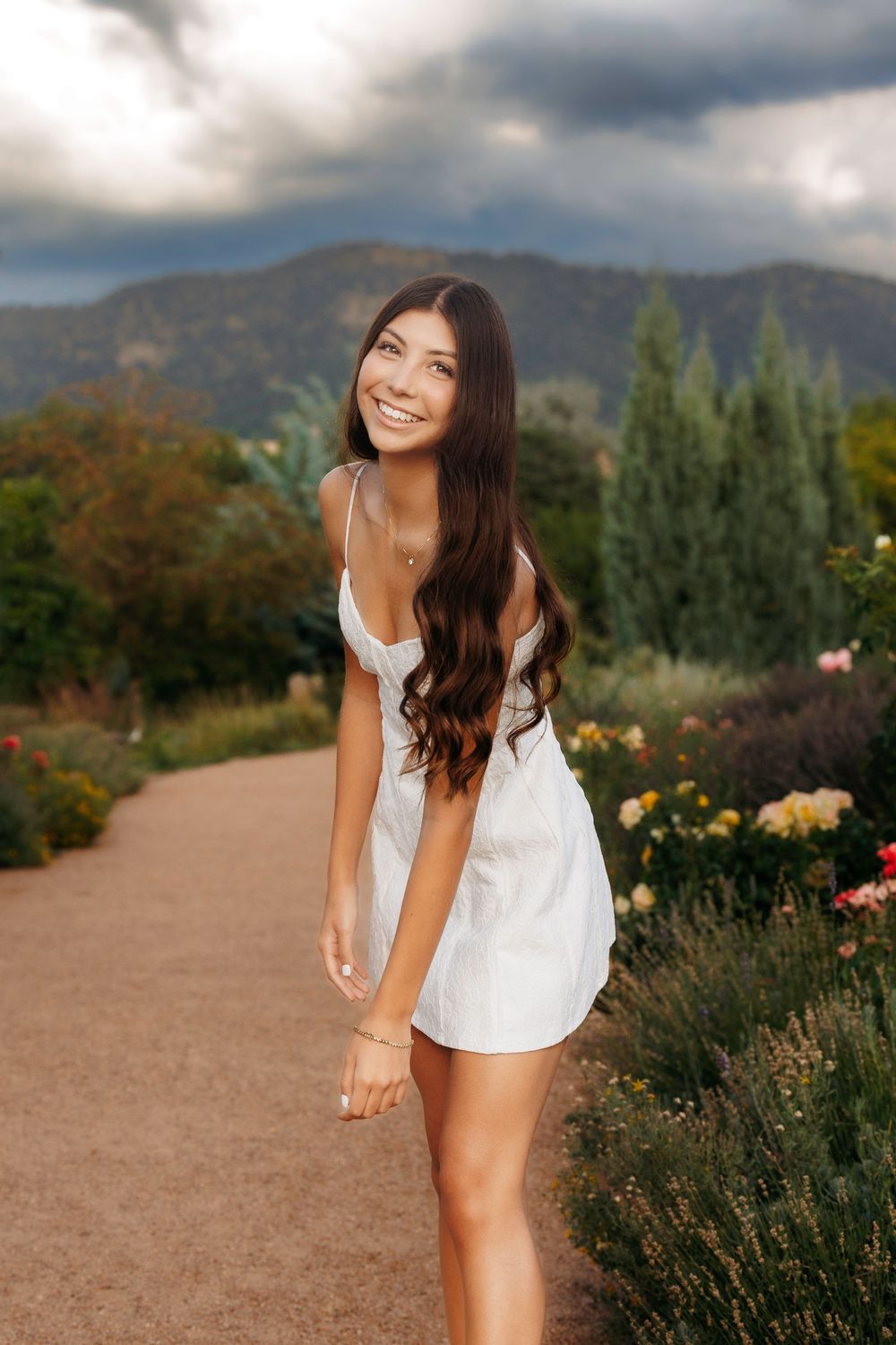 Happy young woman in white dress smiling on a garden path with mountains in the background.