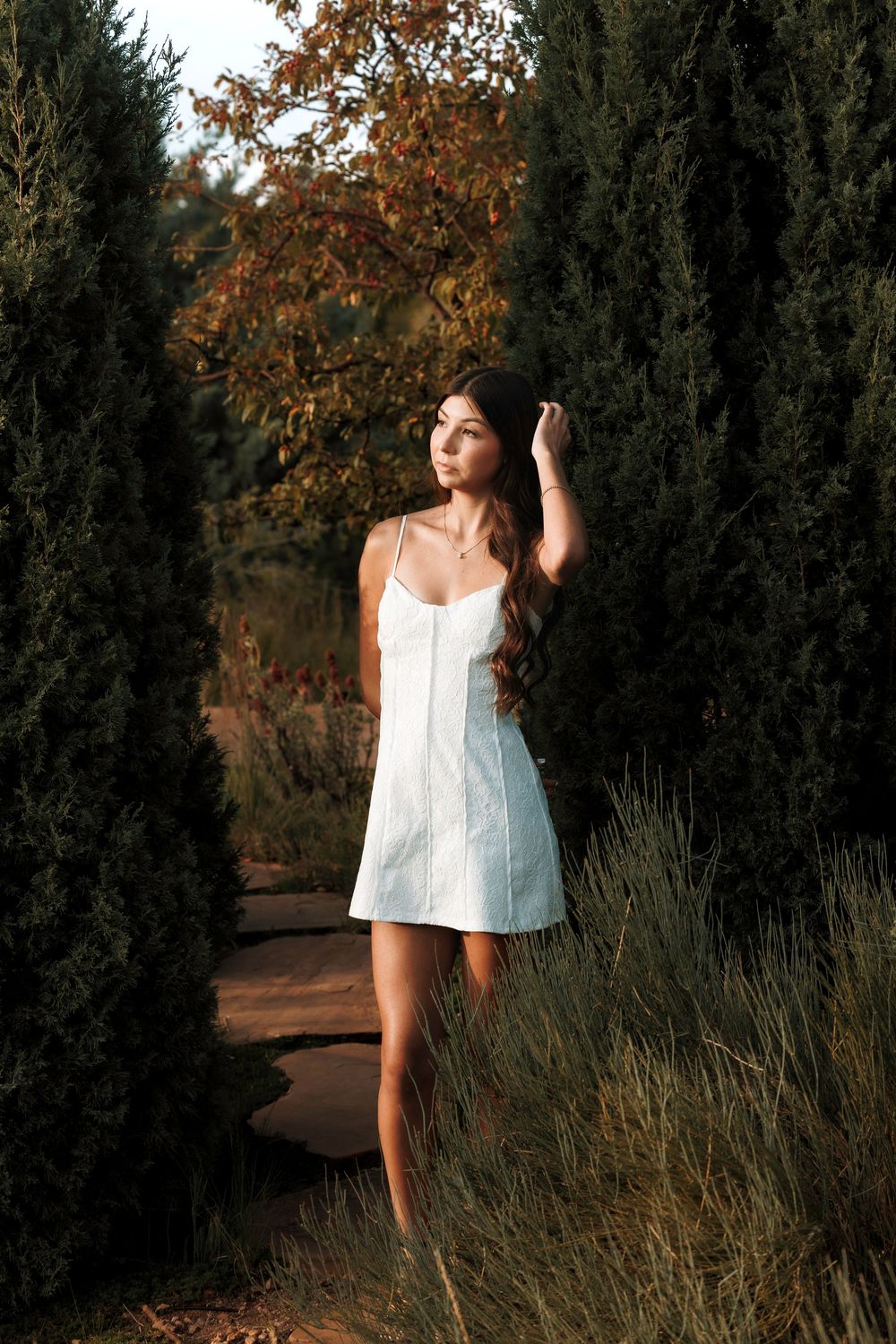 Young woman in white mini dress standing between tall cypress trees in a garden at dusk.