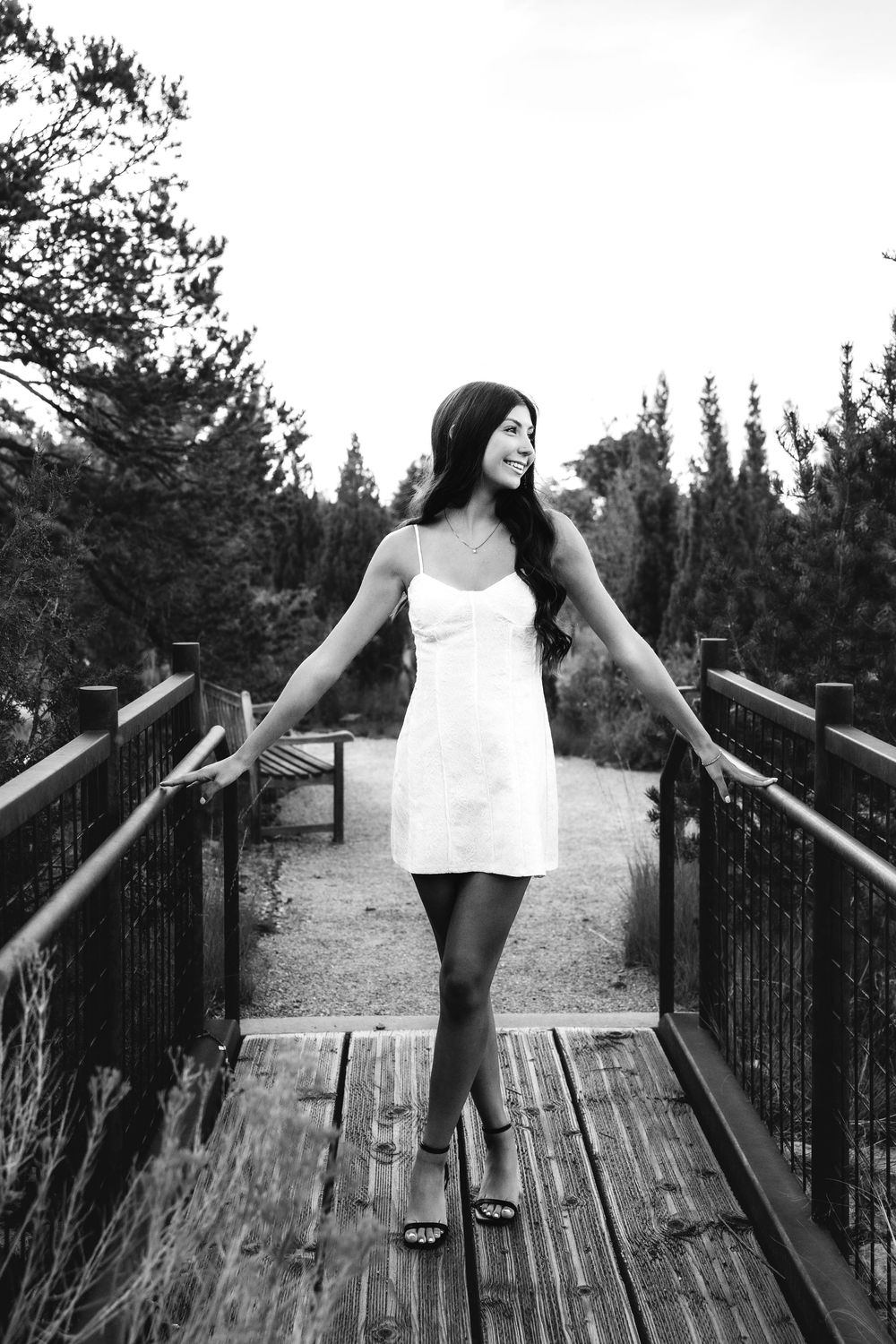 Black and white photo of a young woman in white dress standing on a wooden bridge.