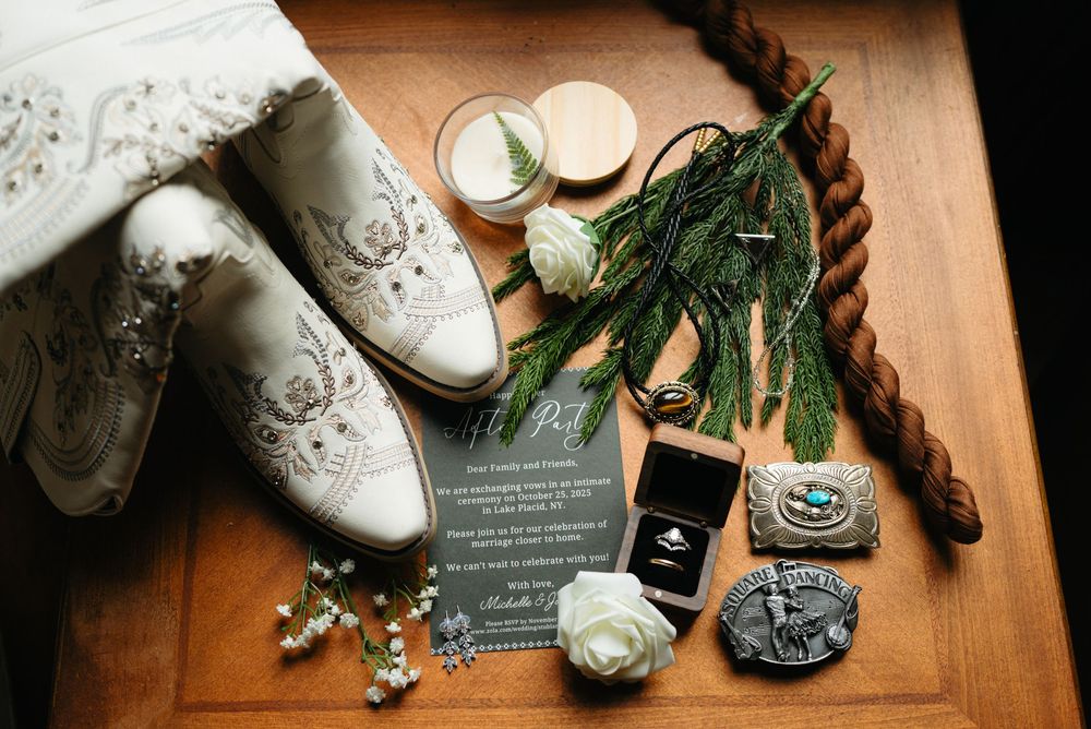 Wedding details including rings, perfume bottle, white shoes, and floral accessories arranged on wooden surface in warm lighting.