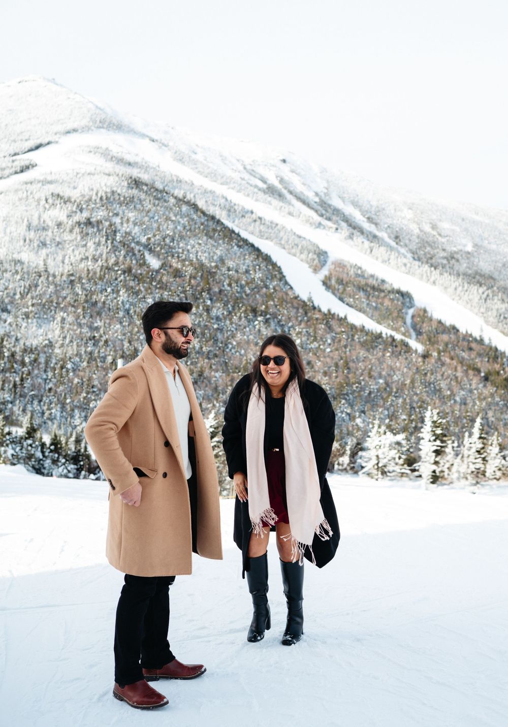 A couple enjoys a romantic winter moment in the snow with mountains and evergreen trees in the background.