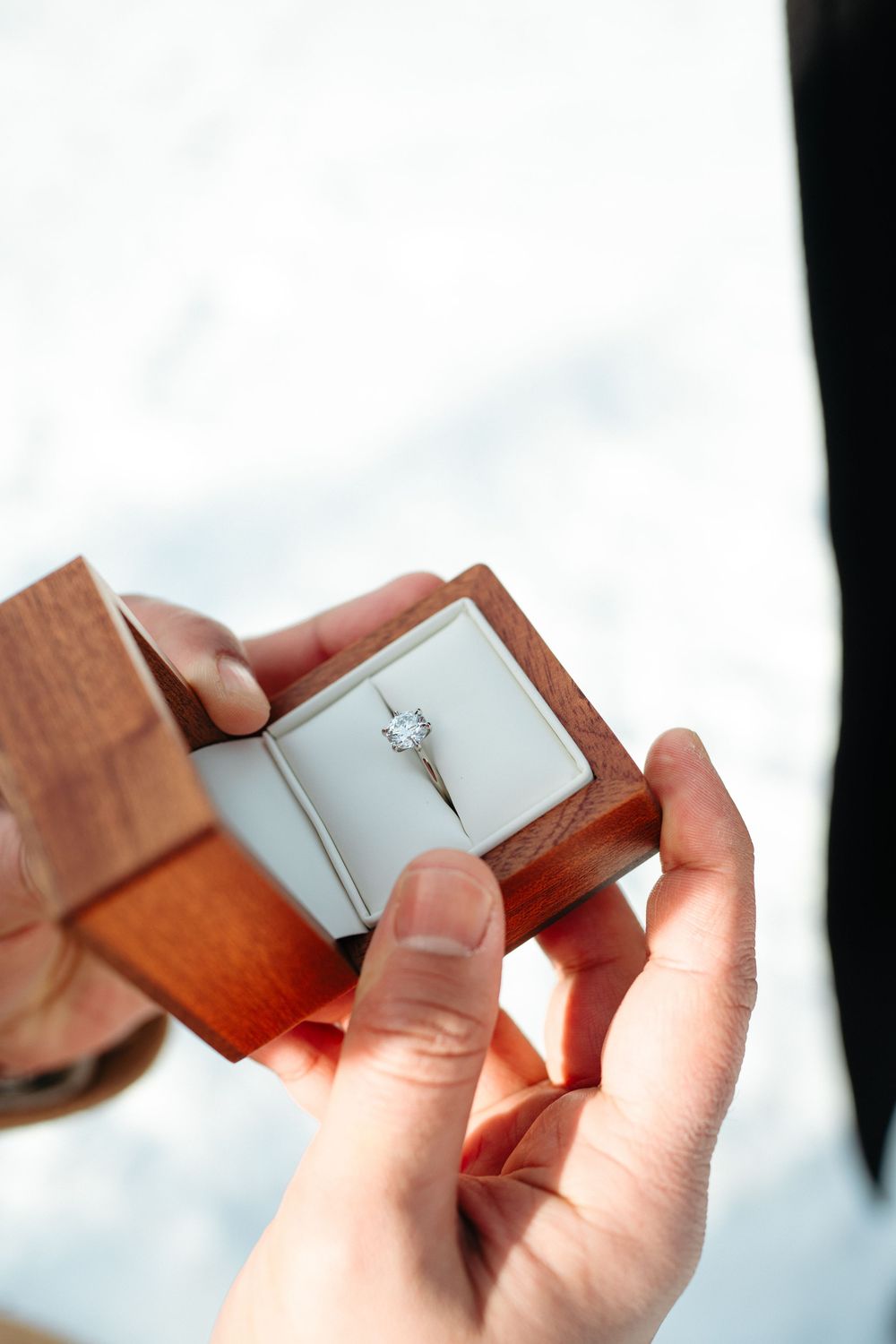 Hands holding a wooden ring box with a solitaire diamond engagement ring against a bright background.