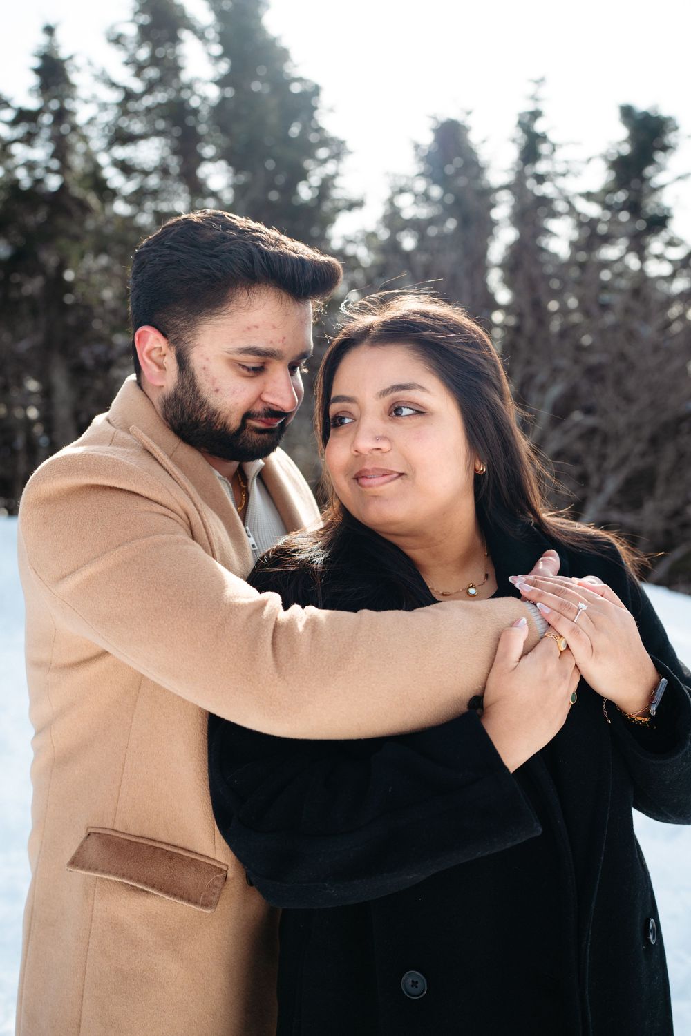 A romantic couple embraces in a snowy winter setting with evergreen trees in the background.
