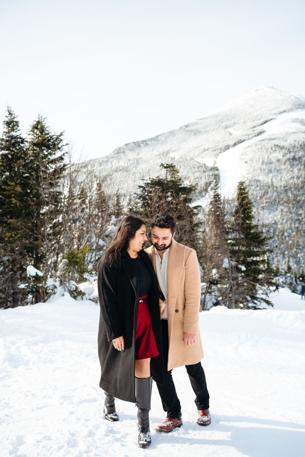 A couple shares a romantic moment in a snowy winter landscape with snow-covered mountains and evergreen trees in the background.