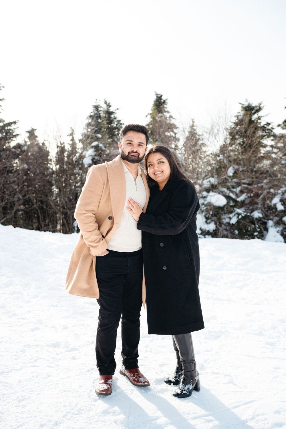 A couple embraces while standing in a snowy winter landscape surrounded by evergreen trees.