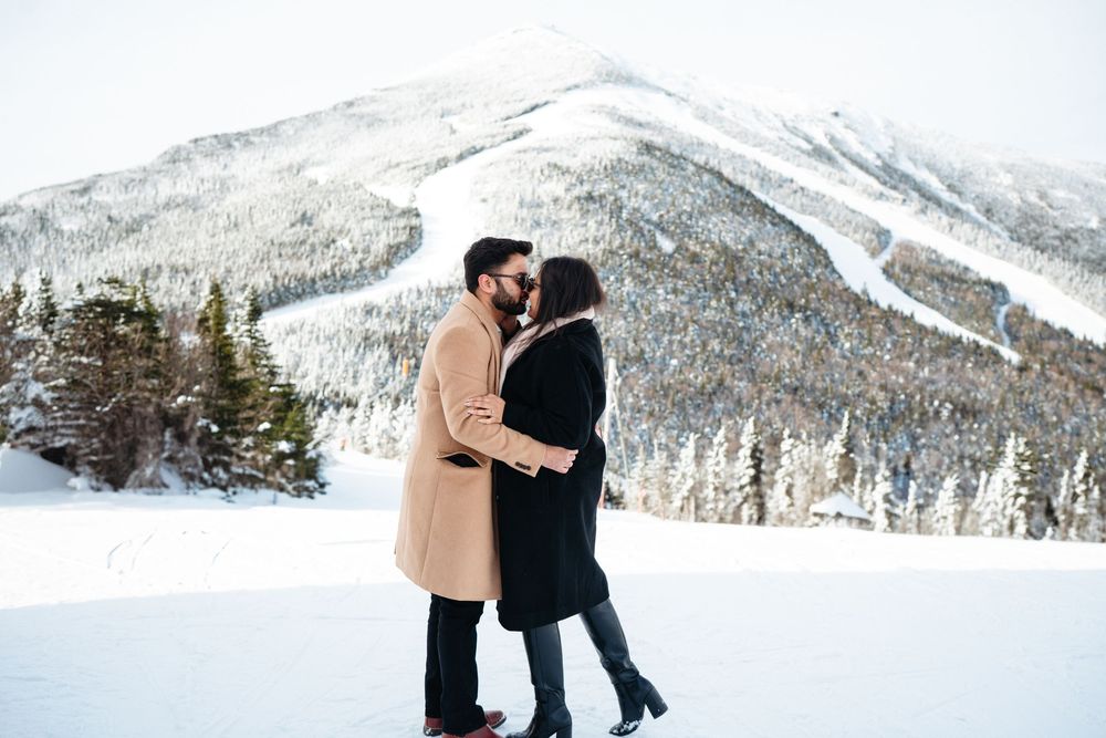 Two people embrace in a romantic winter scene with snow-covered mountains and evergreen trees in the background.