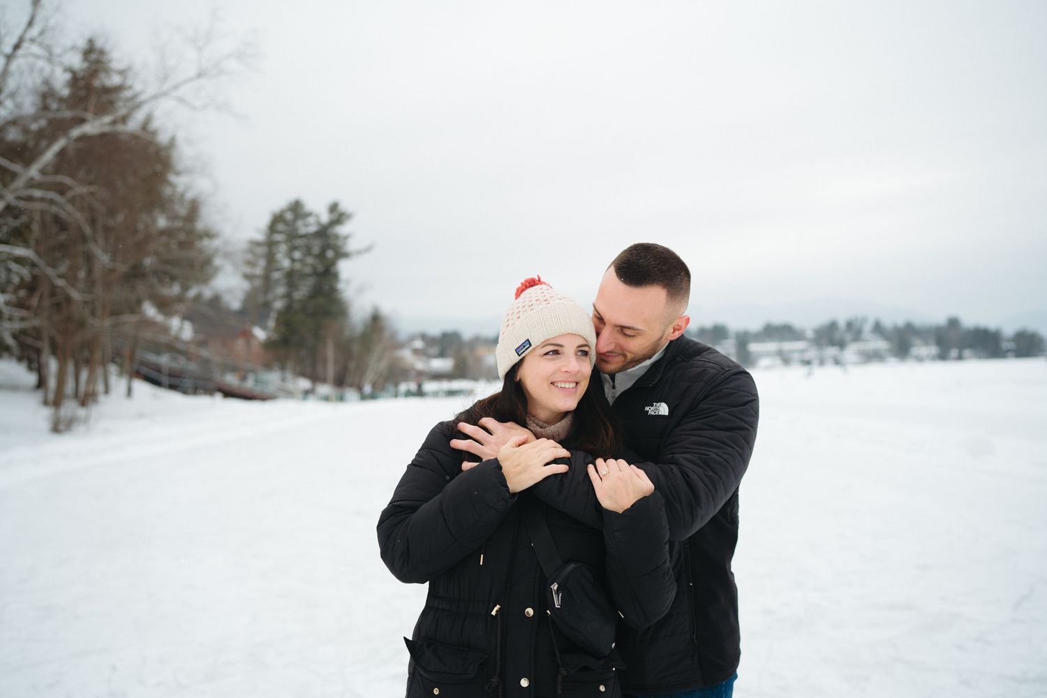 A couple embraces in a romantic moment while standing in a snowy winter landscape.