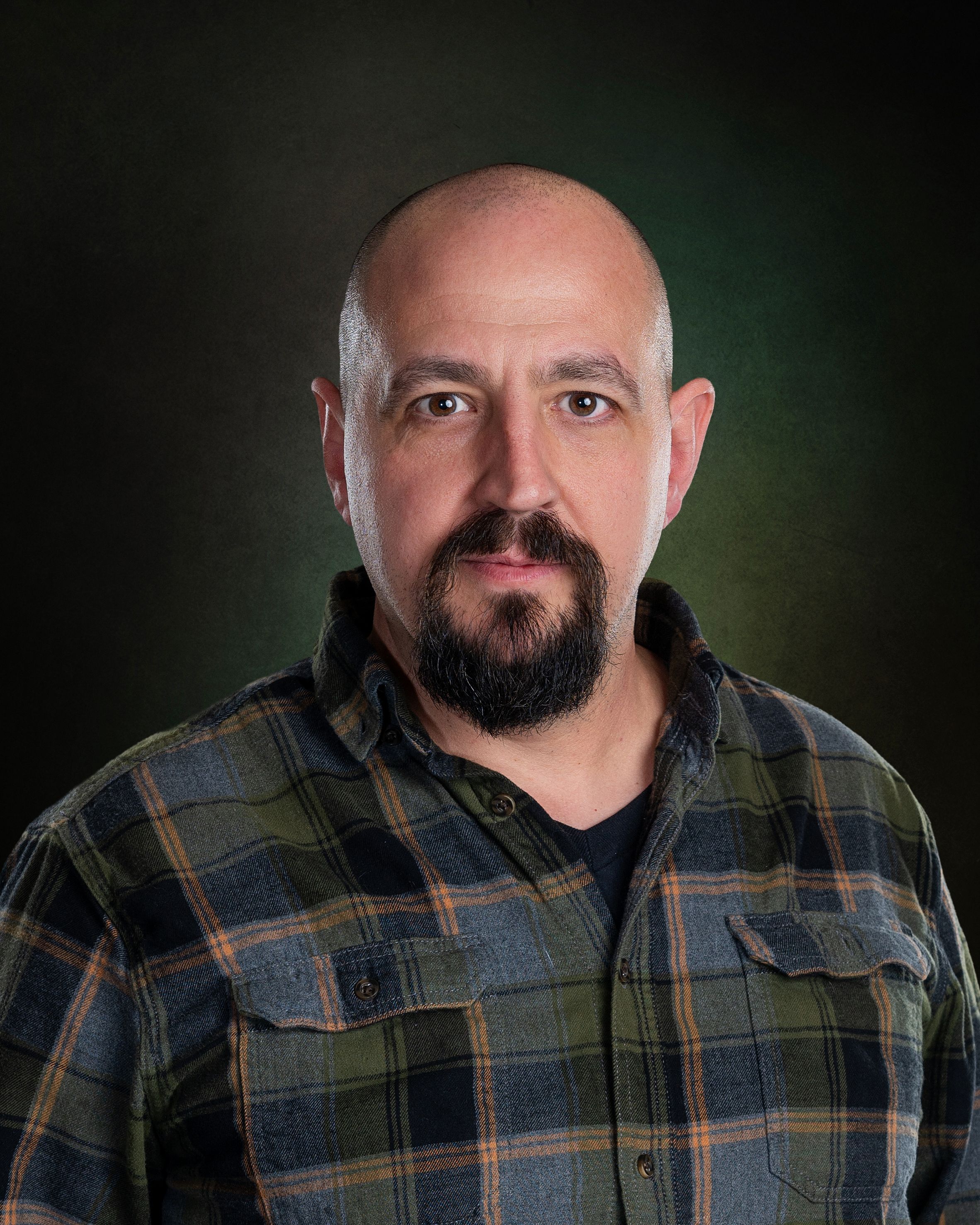 Man in plaid flannel shirt poses against dark studio backdrop.