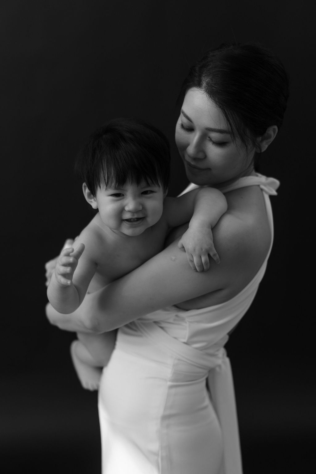 Black and white portrait of a smiling baby held by a woman in a white dress against a dark background.黑白肖像照，一位身穿白色連身裙的女子抱著一個微笑的嬰兒，背景是深色的。