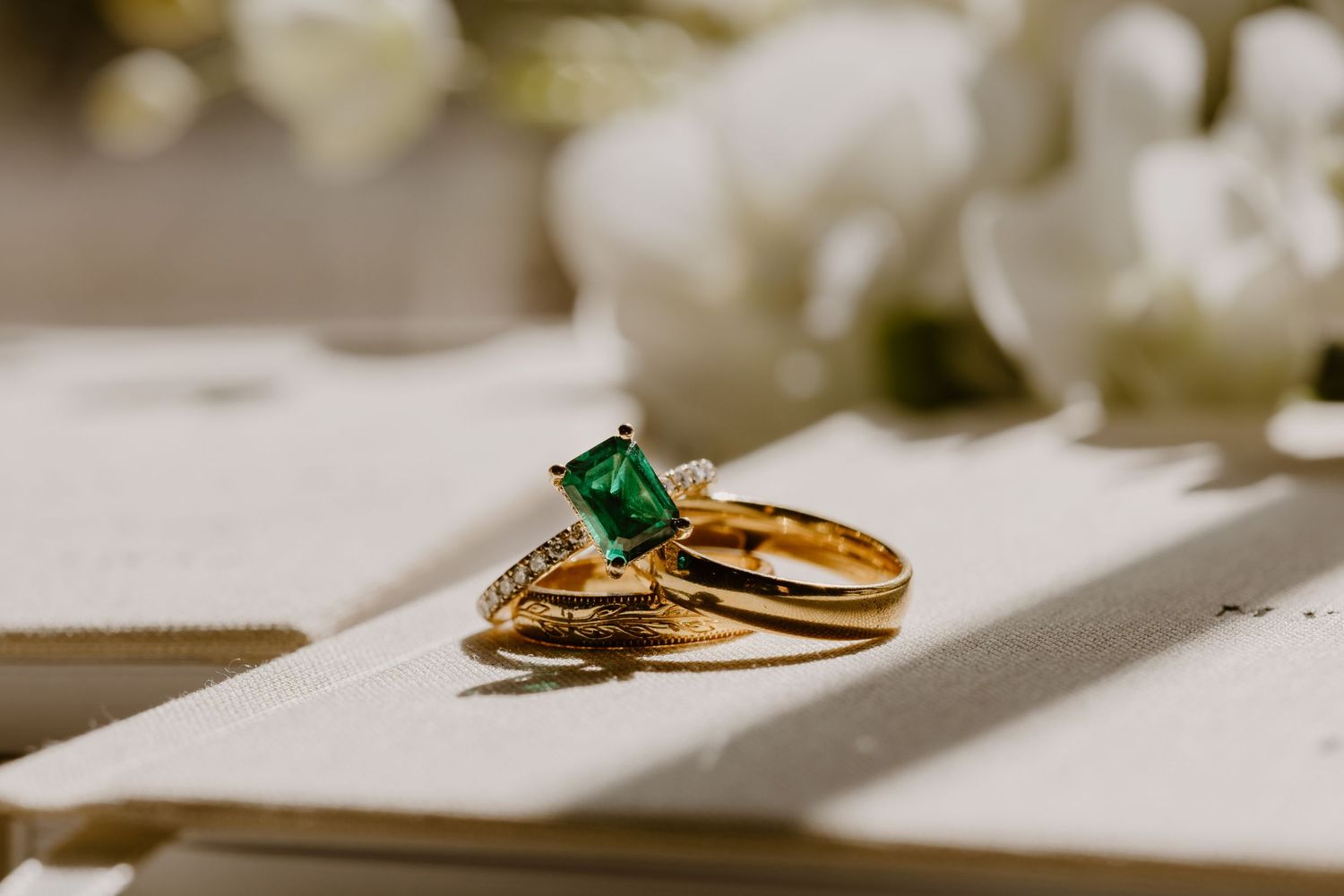 Close-up of emerald cut green engagement ring with gold wedding bands on soft linen with floral background.