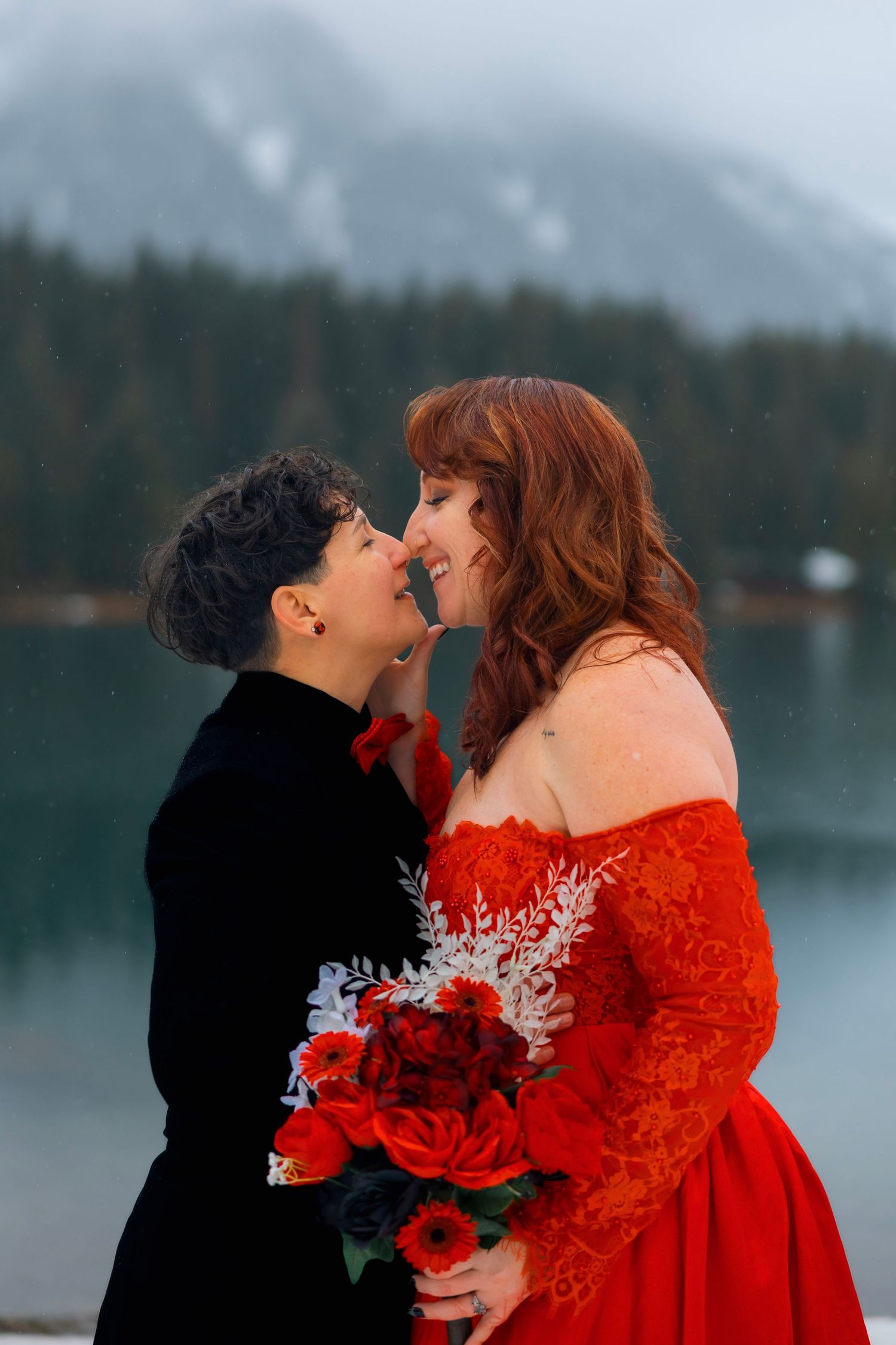 A couple in red and black attire share a clear umbrella in a snowy winter landscape by a lake.