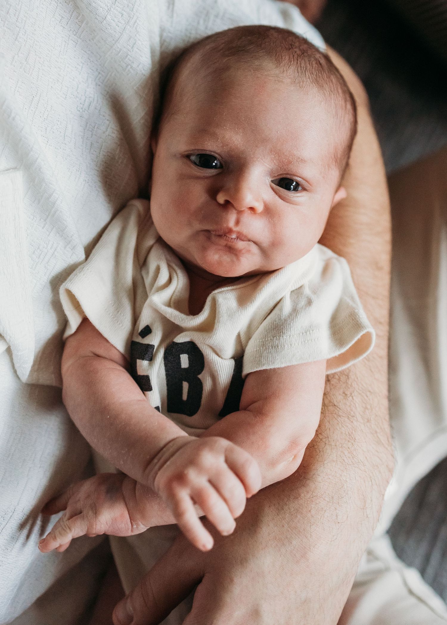 Newborn baby wearing white onesie with text lies on soft white blanket while being cradled in someone's arms.