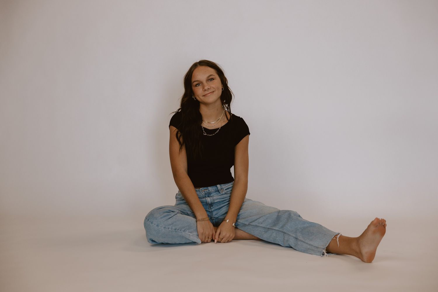 Young brunette woman in black t-shirt and light jeans sitting cross-legged on white studio floor.