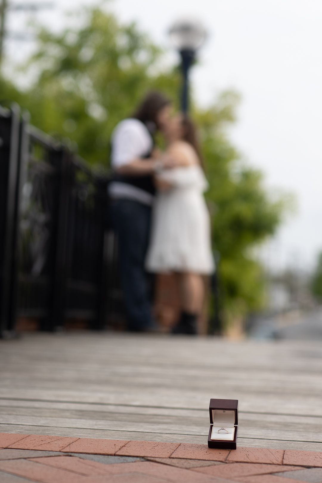 An engagement ring box sits in foreground while a couple embraces on a wooden deck in soft focus background.