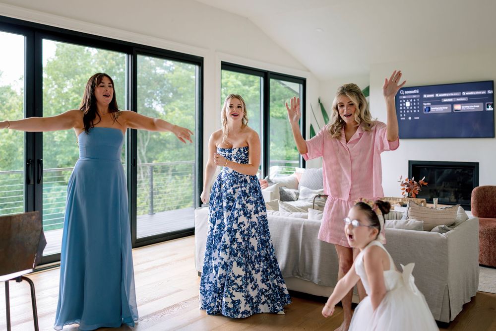 People in formal dresses gather in a modern living room with large windows overlooking greenery.