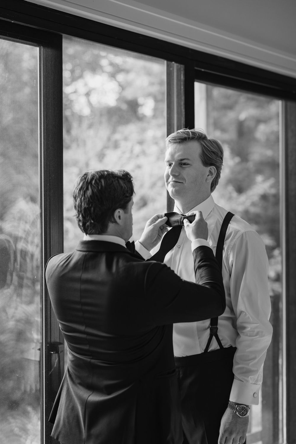 Two people in formal attire stand by large windows in black and white photograph series.