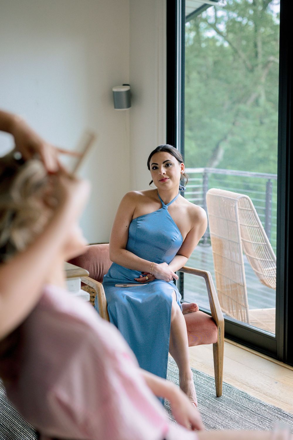 A woman in a blue dress sits comfortably in a rattan chair by large windows overlooking greenery.