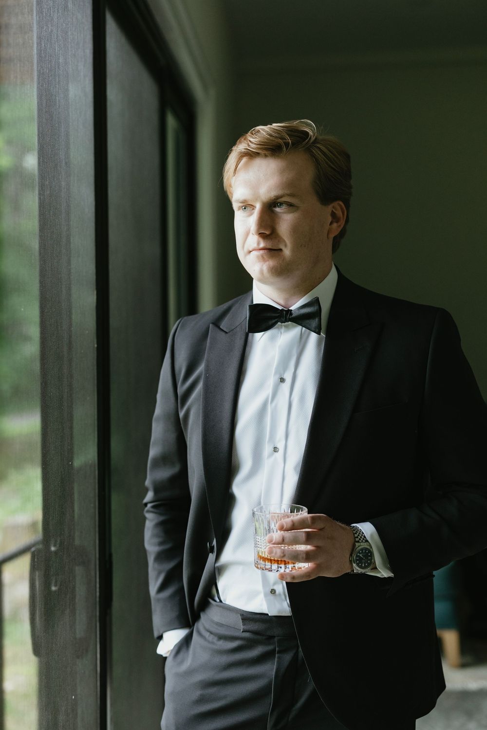 A groom in a classic black tuxedo and white bow tie holds a drink while standing in a rustic covered porch.