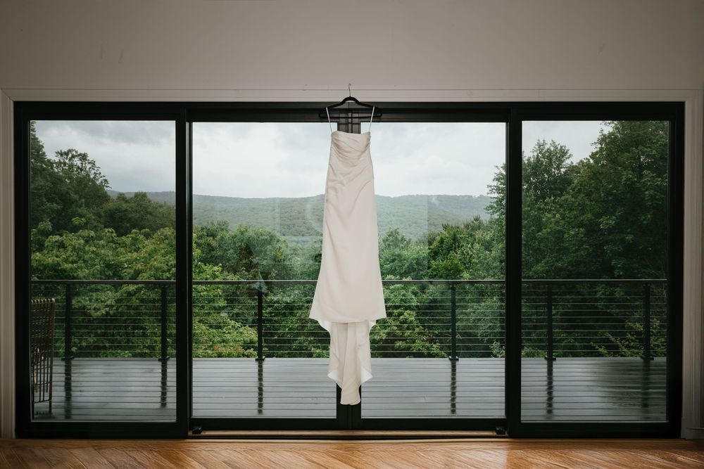 A series of panoramic windows in a modern room overlooks a lush green mountain landscape with a white dress hanging.