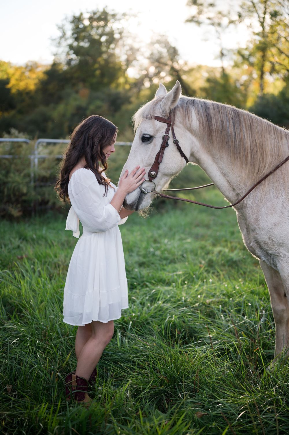 Person in white dress interacting with white horse in sunlit meadow.