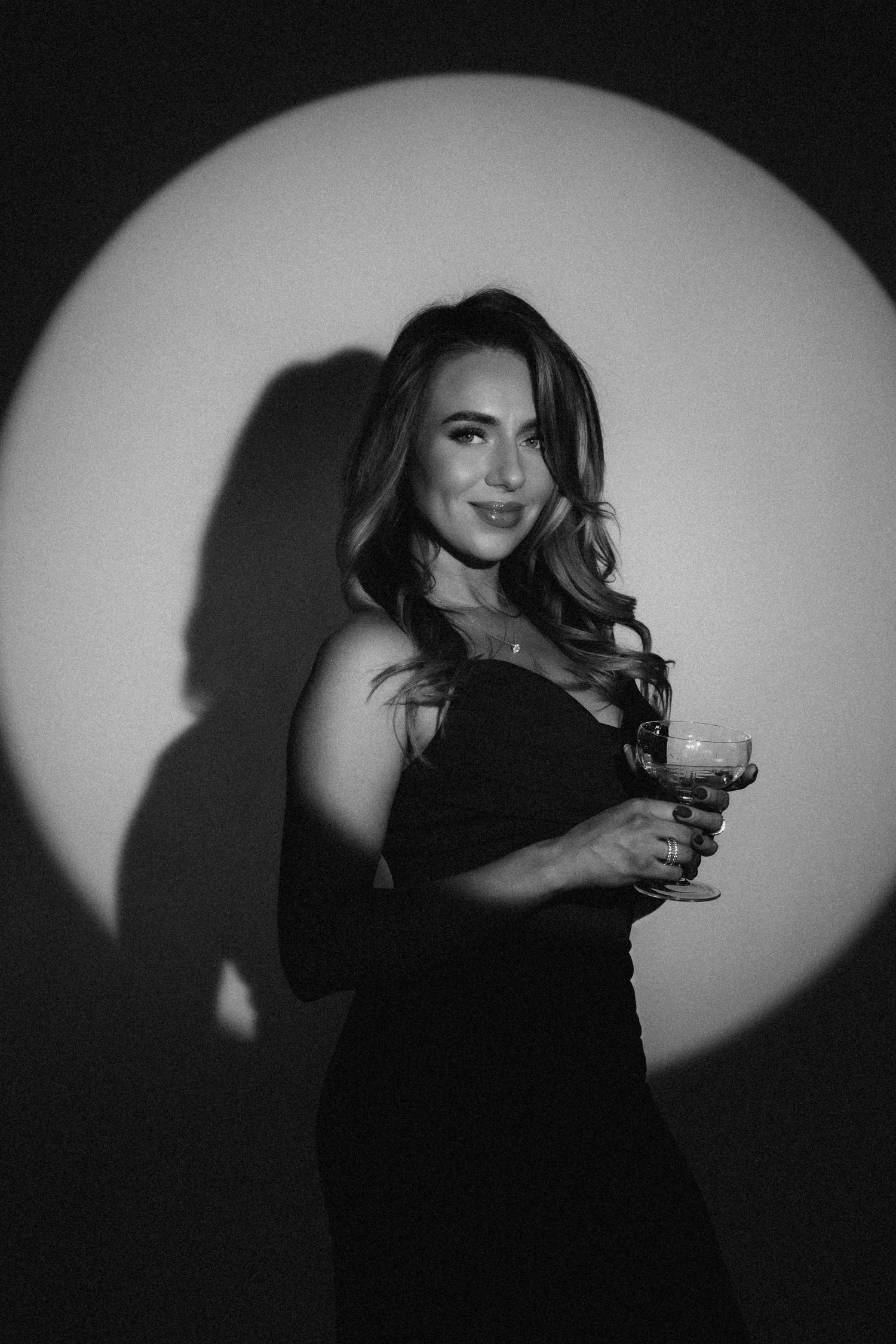 Black and white photo of elegant woman in black dress holding cocktail glass with dramatic spotlight background.