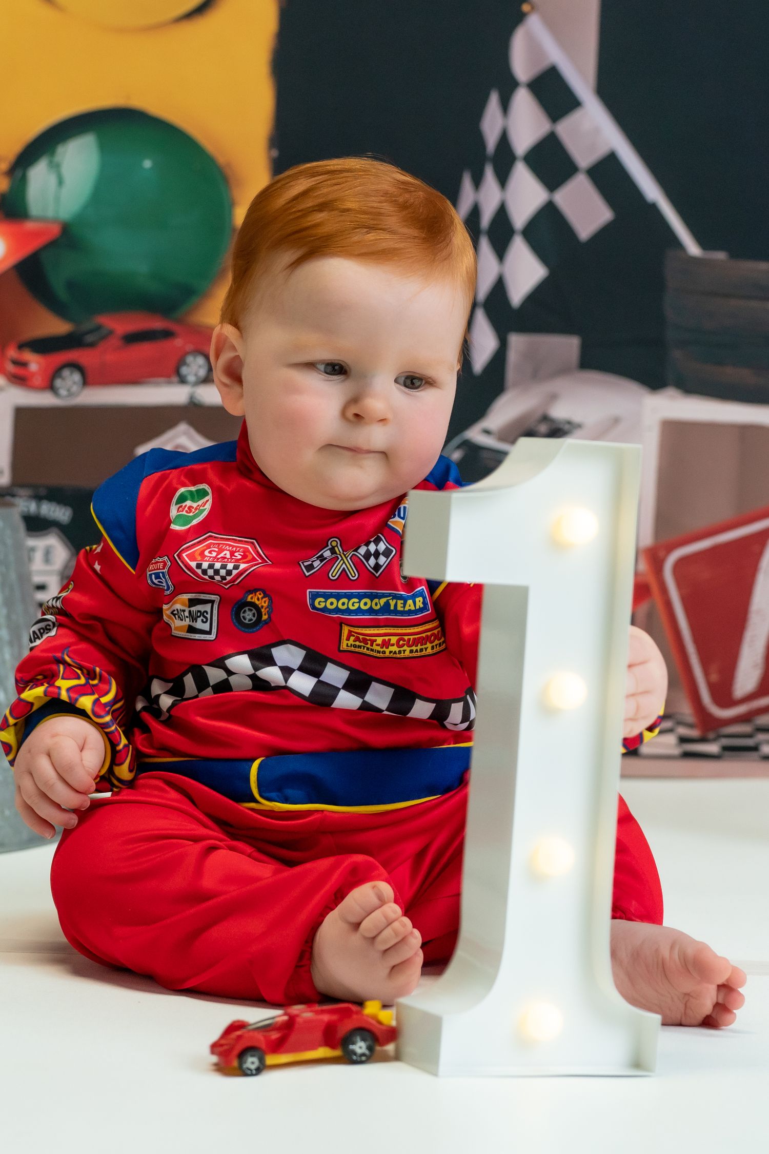 Baby in red racing outfit sits beside number one decoration and toy car with racing-themed decor in background.