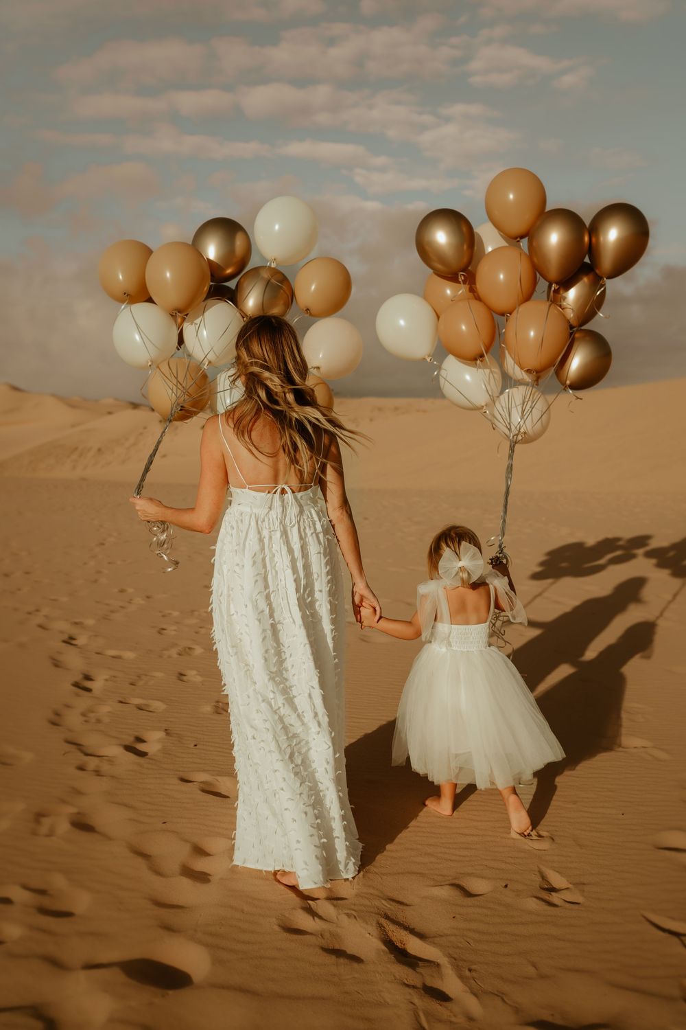 A mother and daughter sydney photoshoot hold golden balloons while walking through desert sand dunes at sunset.