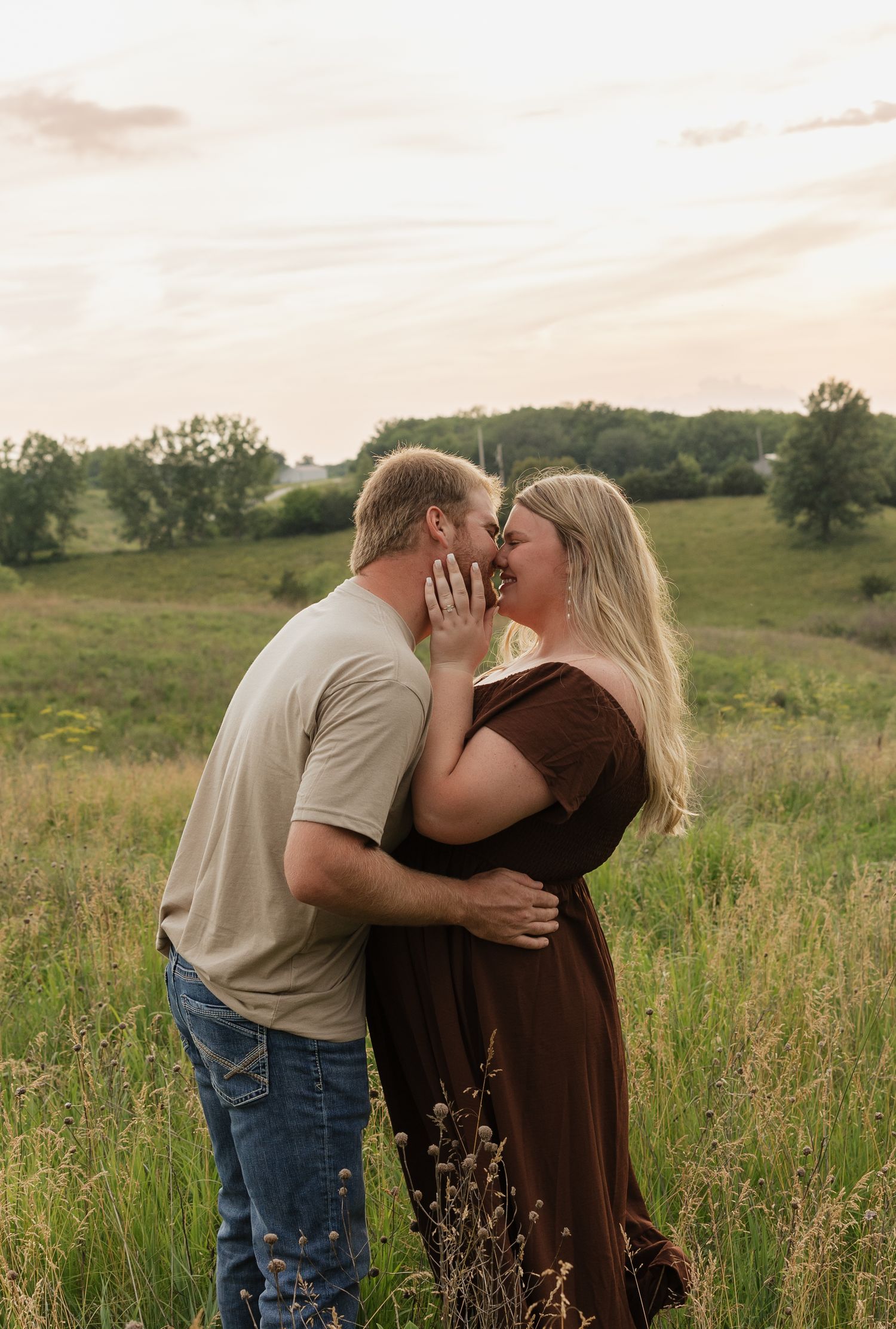 A romantic couple shares an intimate moment together in a sunlit meadow with tall grass and trees in the background.