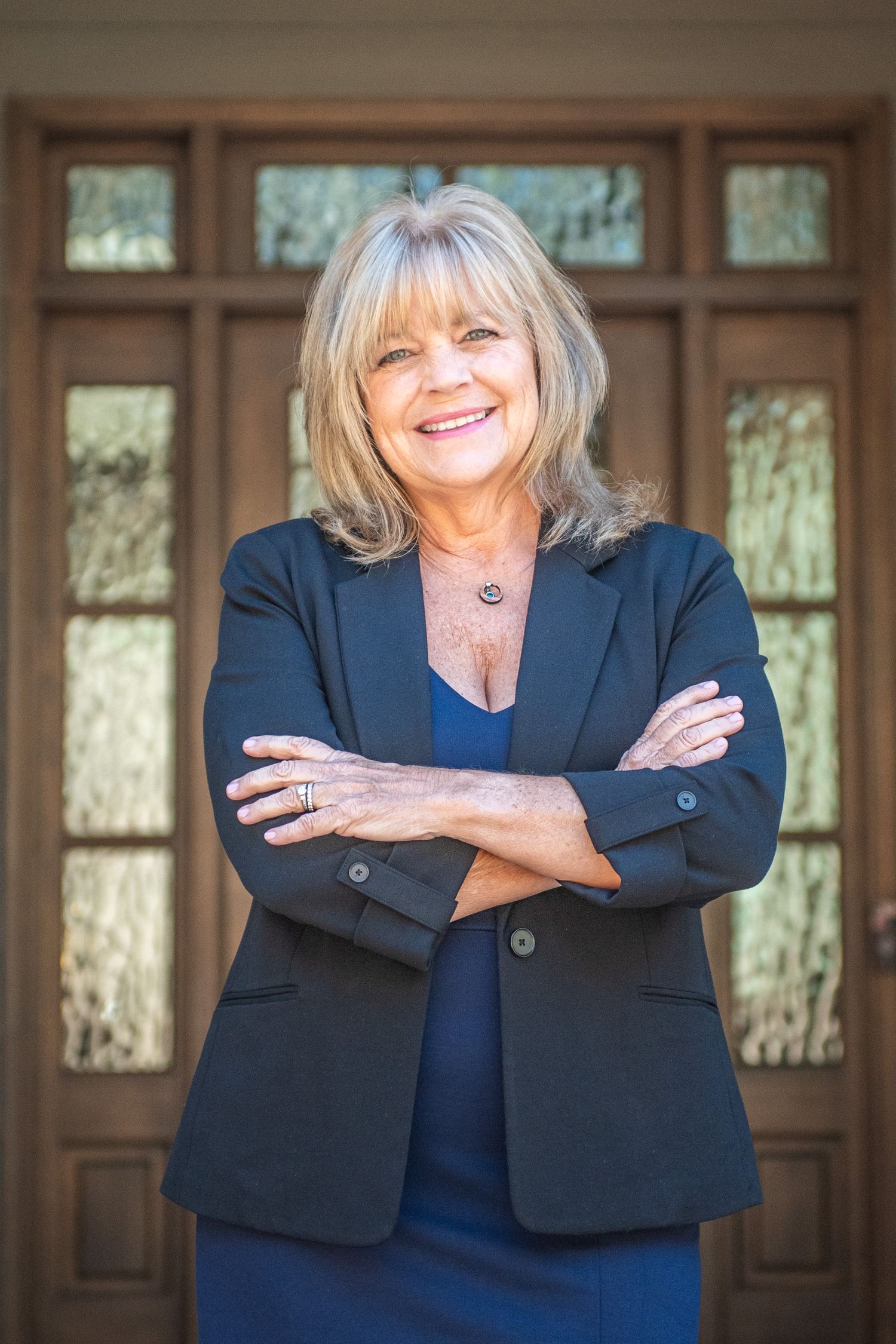 Professional portrait of a person in a navy blazer and dress standing confidently with arms crossed in front of wooden doors.