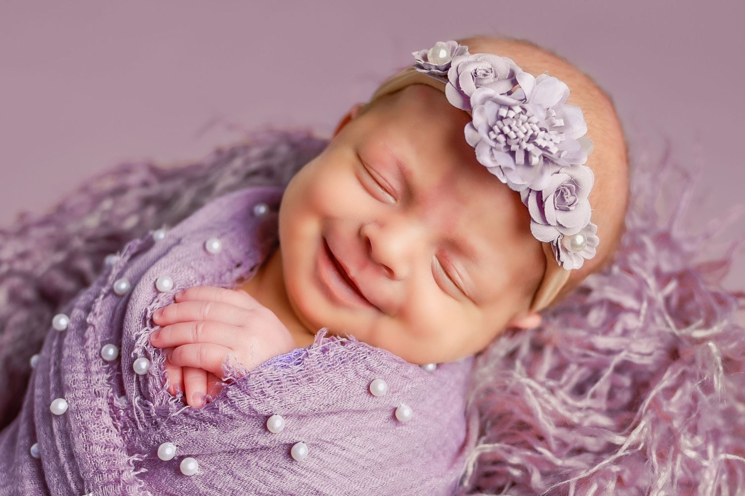 Newborn portrait photography of a baby girl wrapped in purple fabric smiling in her sleep.