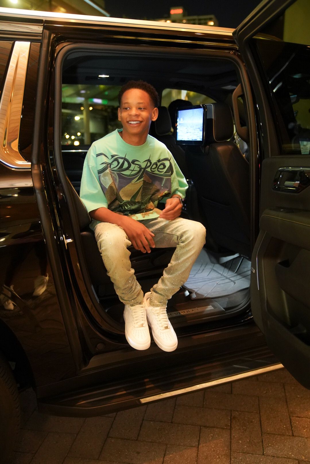 Young person in a mint green shirt and white sneakers sits in the doorway of a black SUV at night.