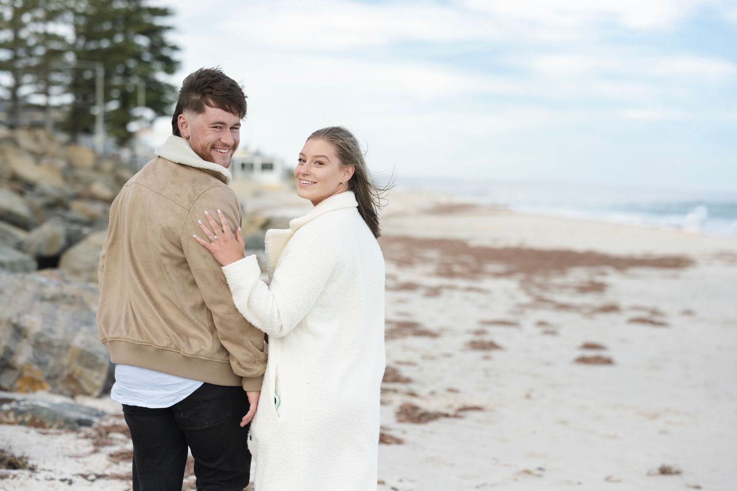 Couple embracing on a sandy beach in winter coats, smiling over their shoulders with rocky shoreline behind them.