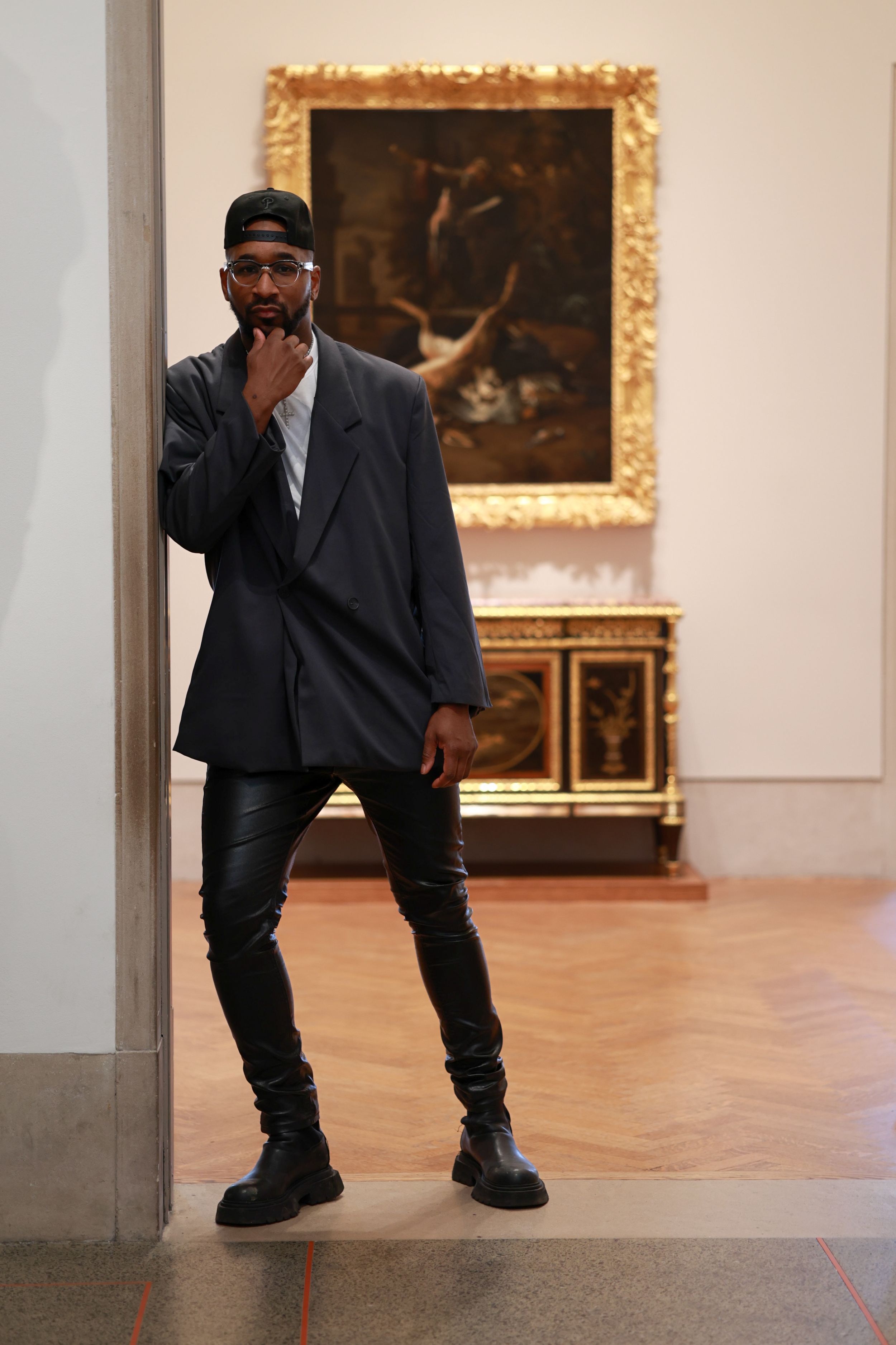 A person in dark clothing crouches and poses dramatically in an art gallery with framed paintings on the walls.