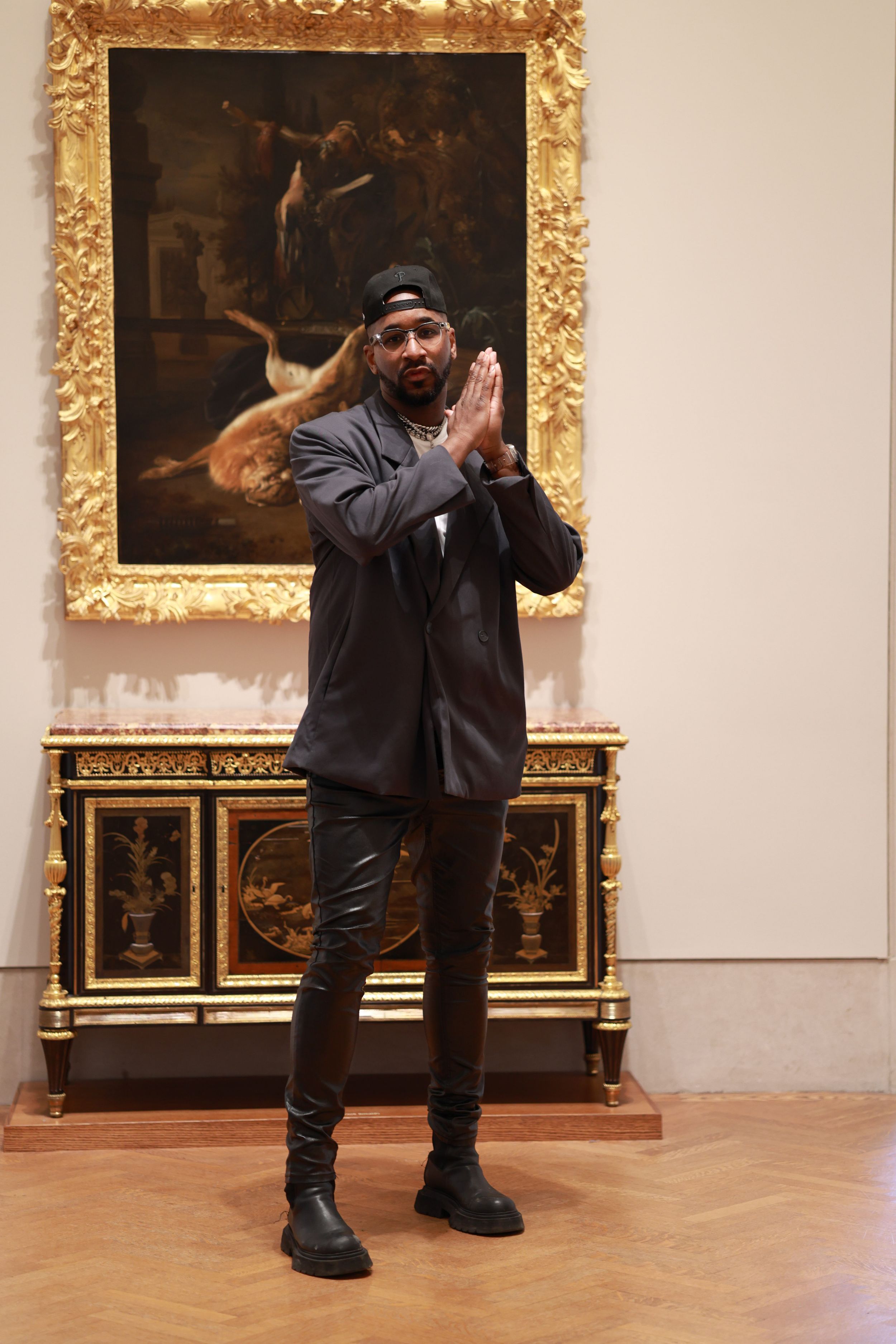 A person in dark clothing strikes a playful pose in front of classical artwork at an ornate museum gallery.