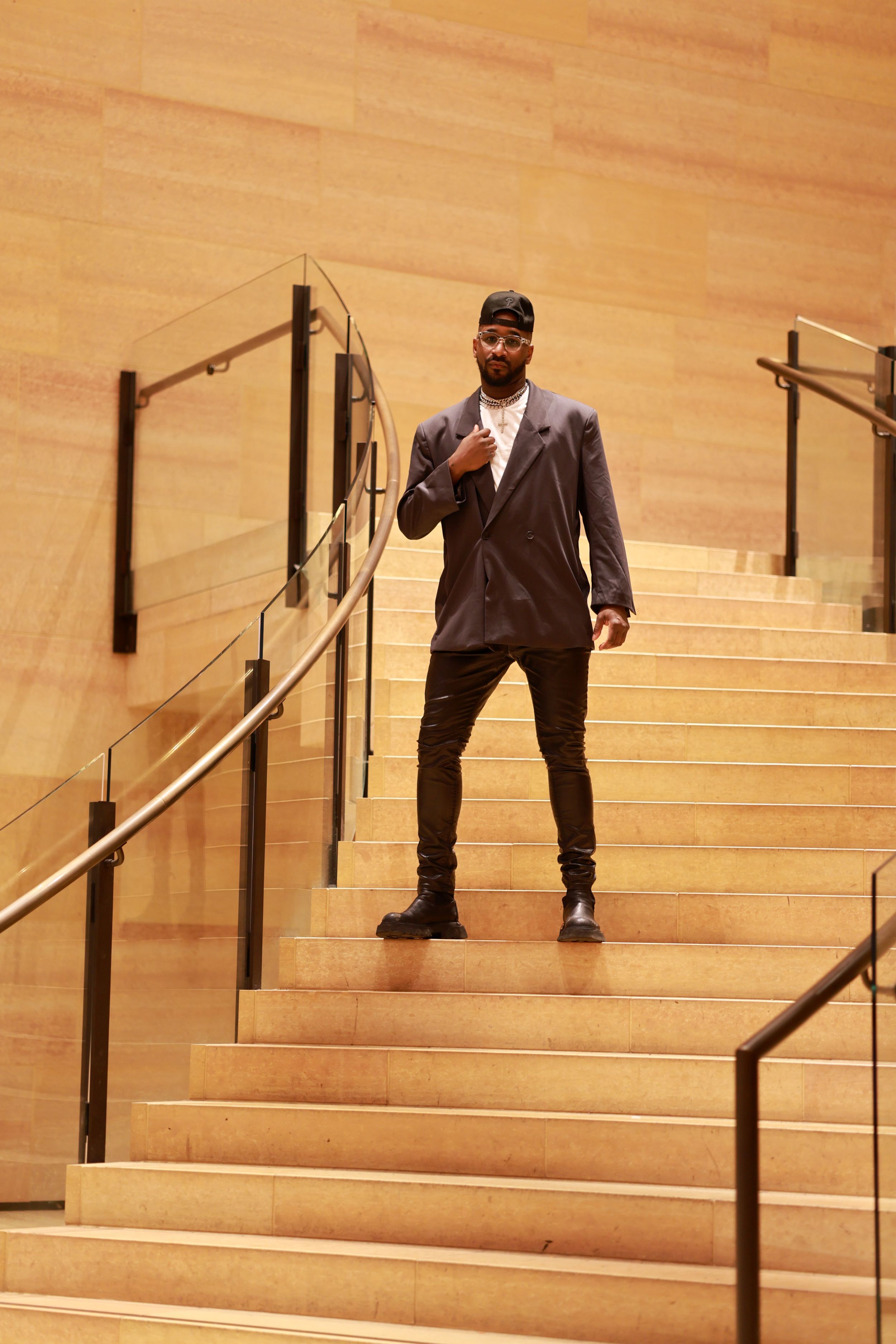 Someone wearing a baseball cap and dark jacket poses confidently on wooden stairs with metal handrails in an indoor setting.