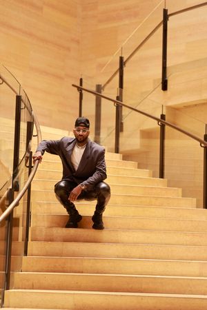 A person crouches on wooden stairs with glass railings in an elegant indoor setting.