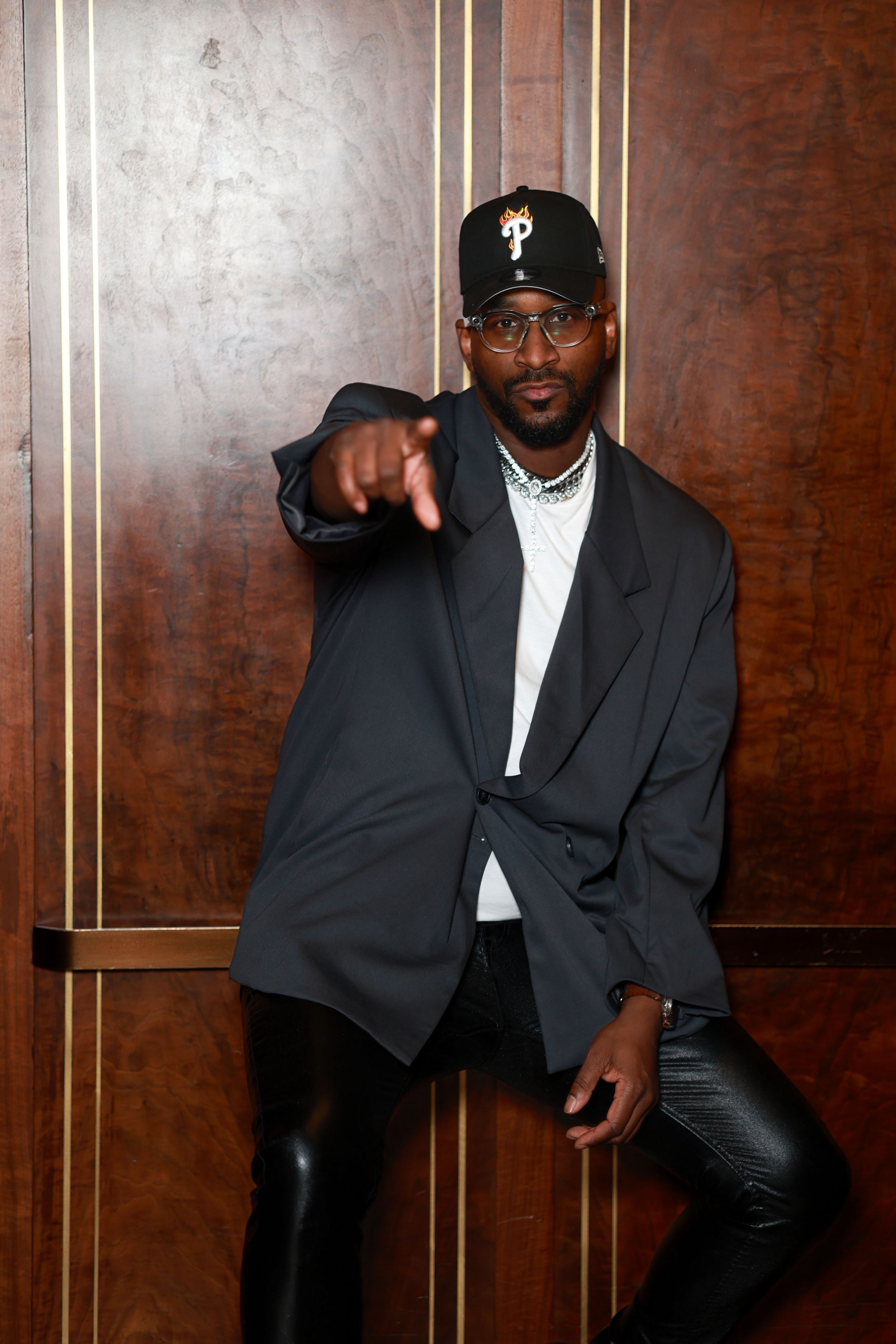 A person wearing glasses, a baseball cap with a Phillies logo, and a dark blazer points at the camera while posing against a wall.