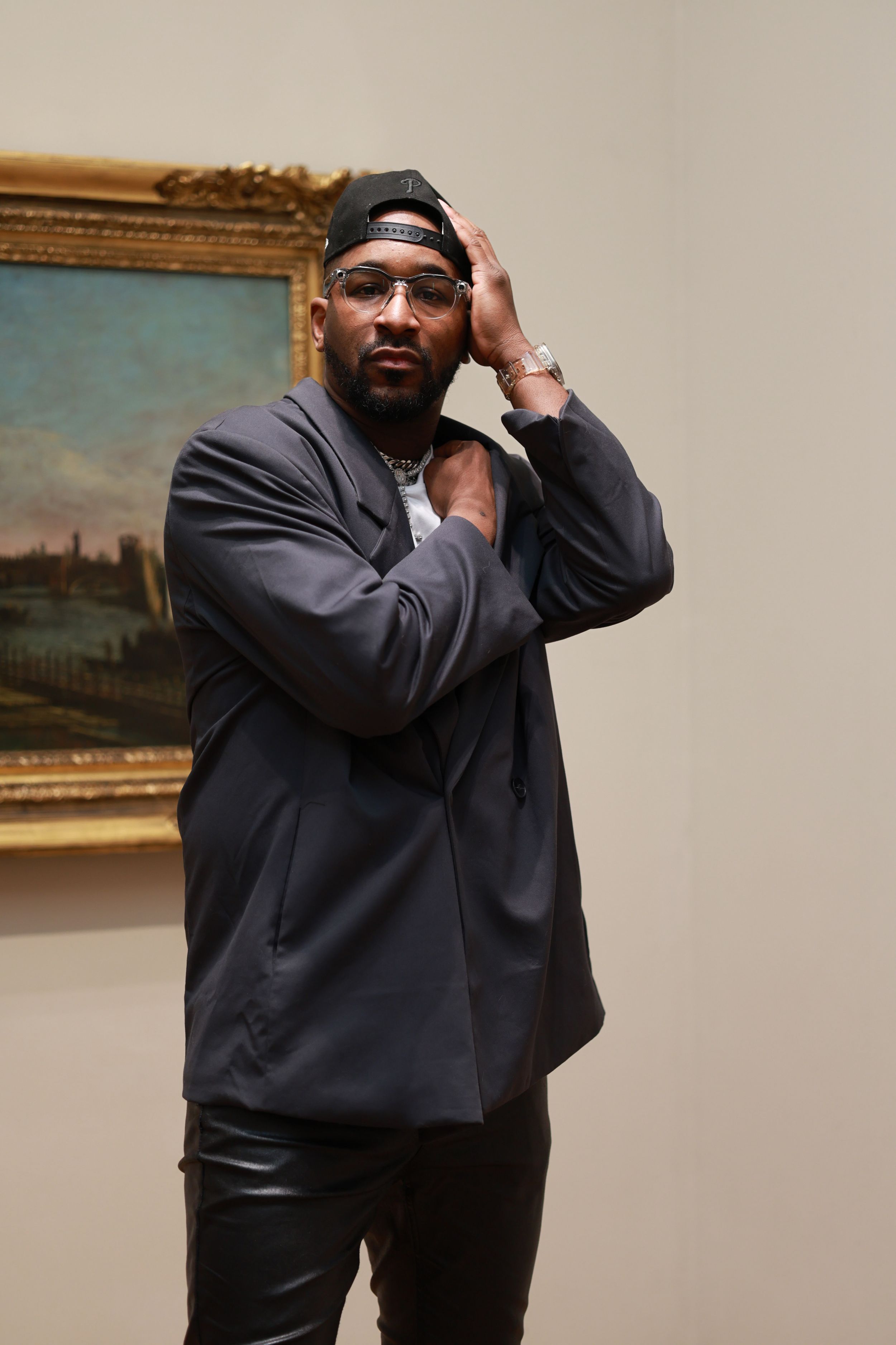 A person wearing a black jacket and backwards cap poses thoughtfully against a wall with framed artwork.