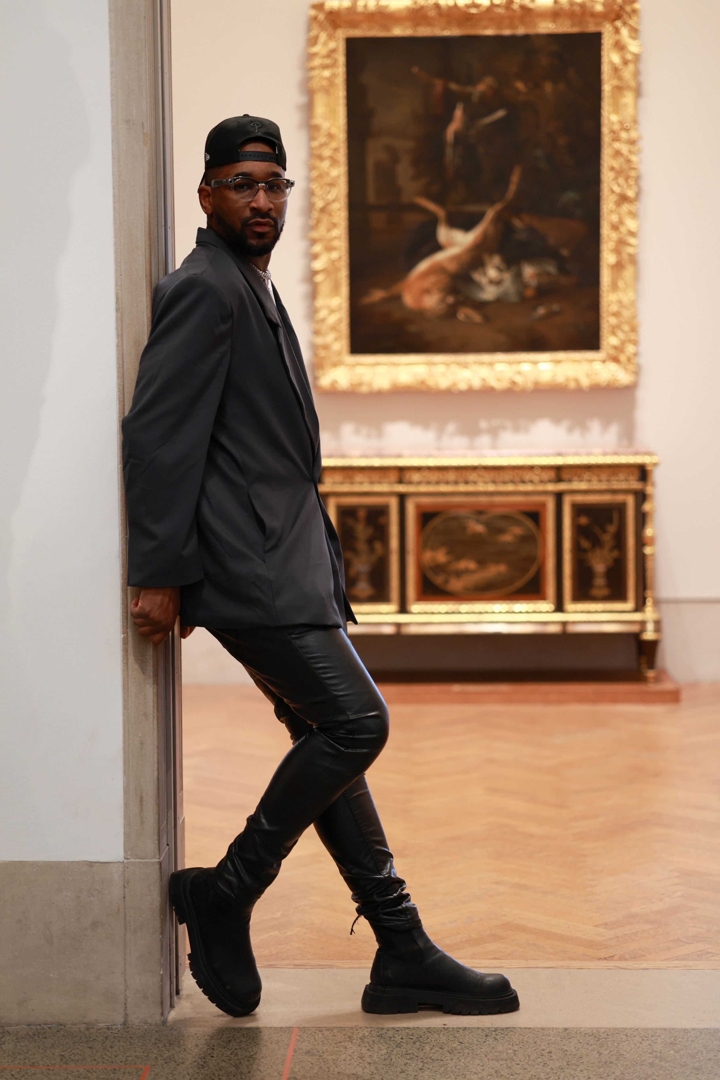A stylishly dressed museum visitor poses against a wall near a classical painting in an elegant gallery setting.