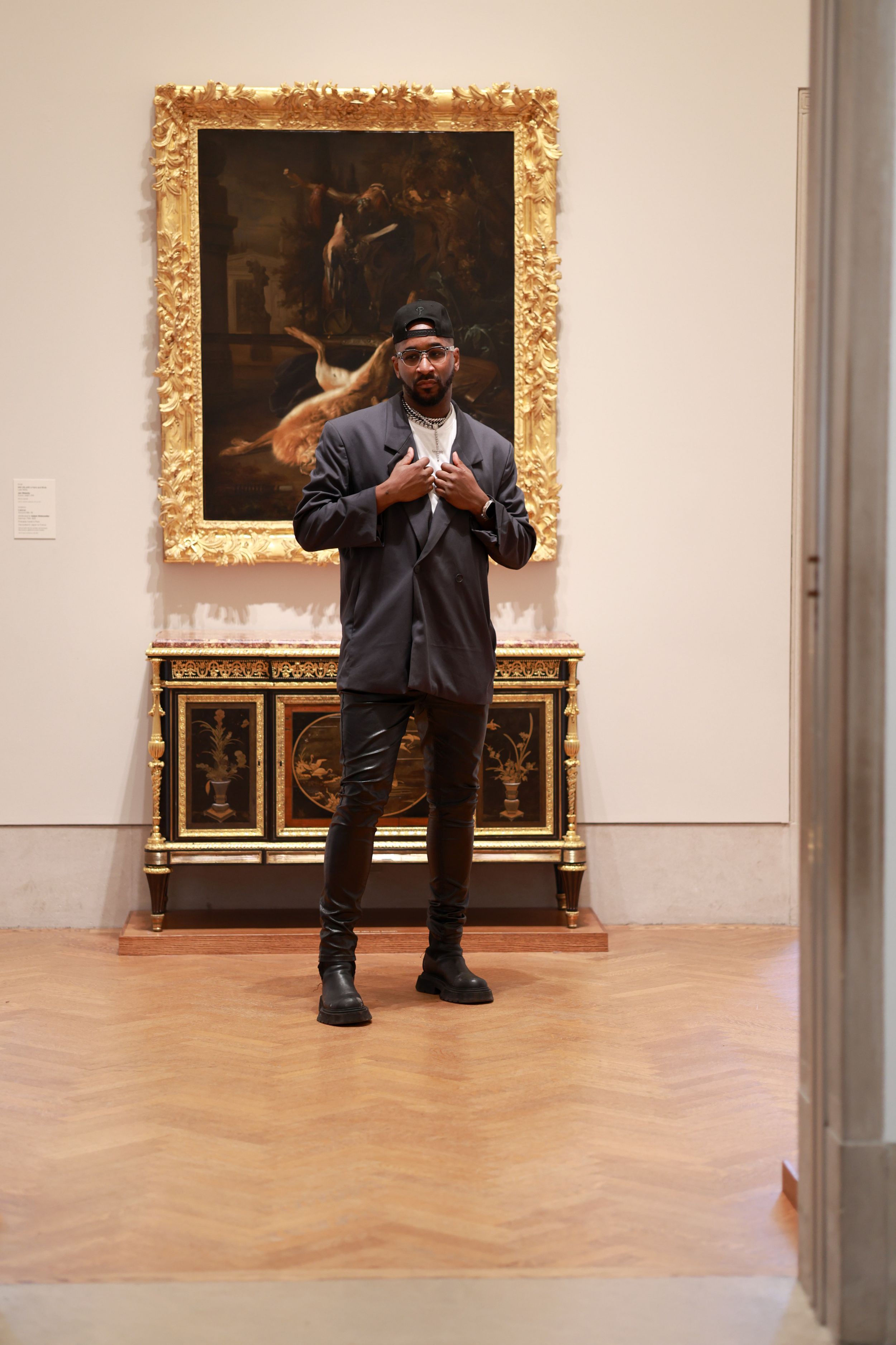 A visitor strikes a pose in front of a classical painting with ornate gold frame in an art museum gallery.