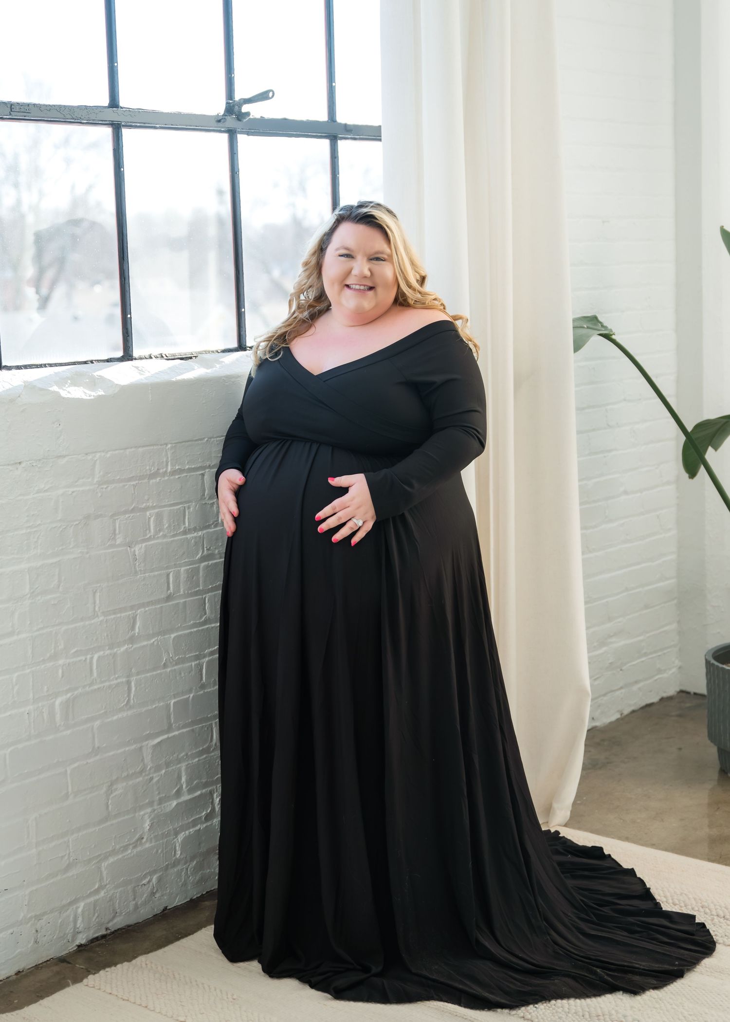 A person in a flowing black dress poses gracefully in a bright studio space with white brick walls and natural light.